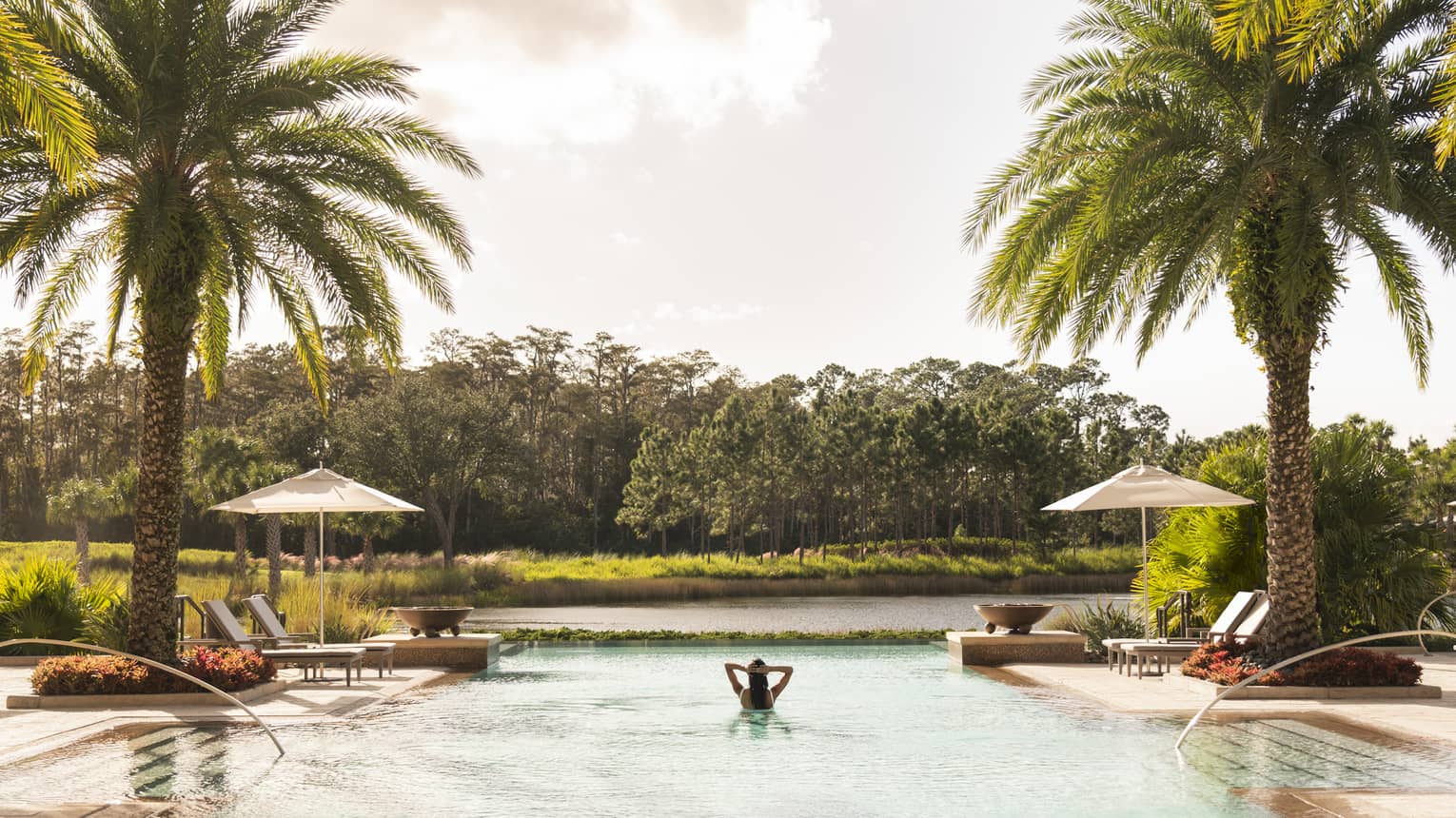 A woman alone in a large pool with lounge chairs and palm trees around it.