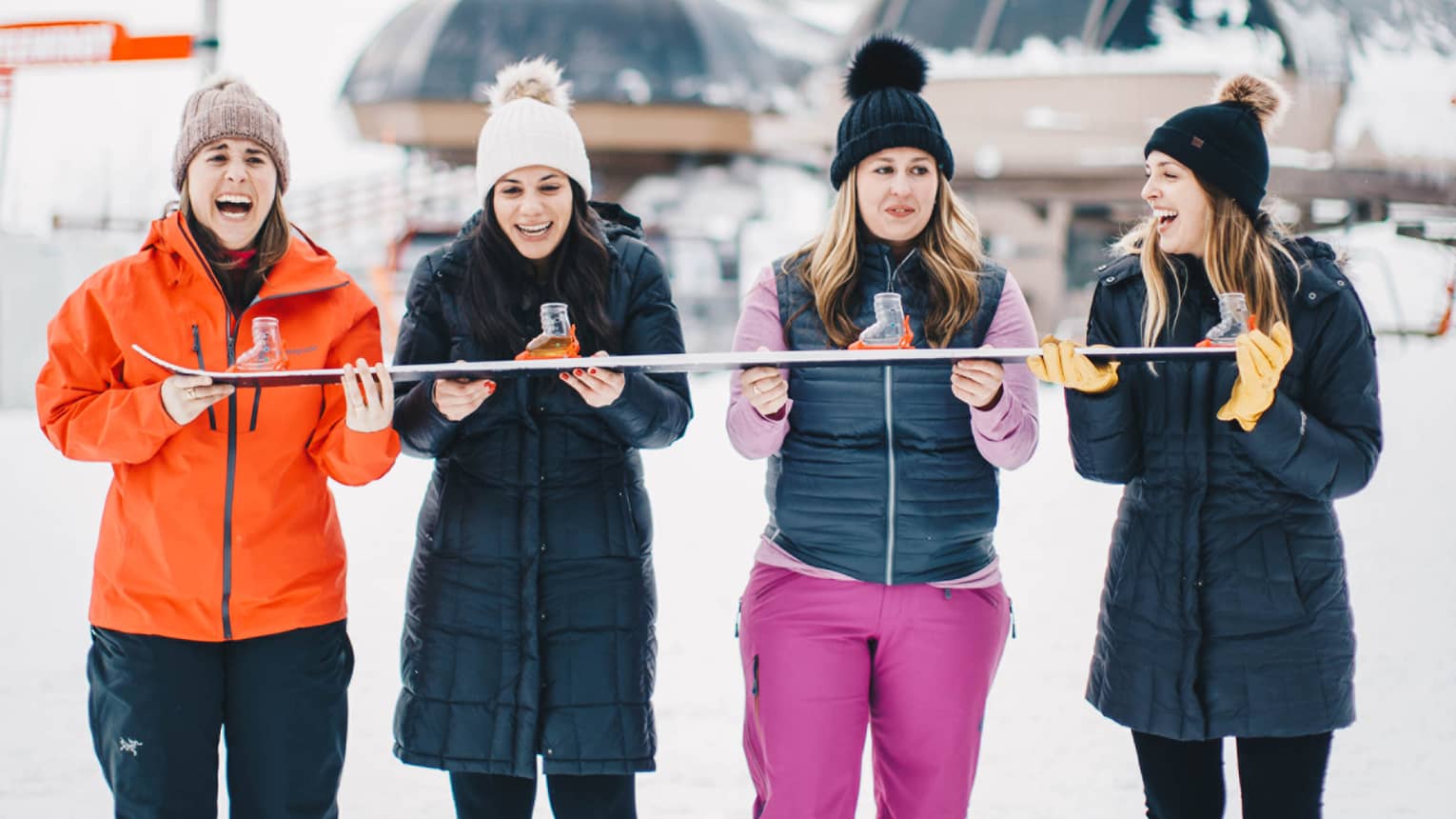 Four people in winter outdoor clothing stand in the snow, holding a ski, with snow-covered mountains in the background