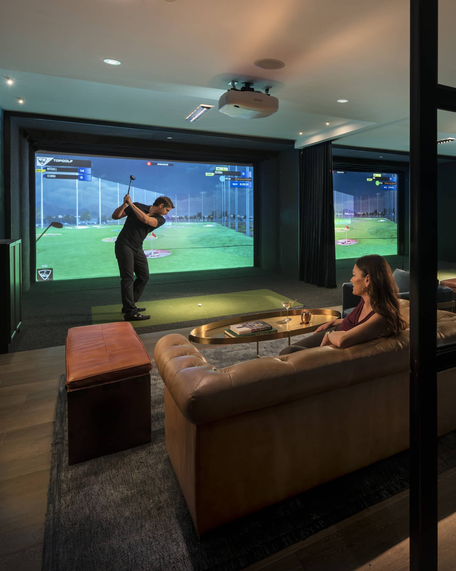 Topgolf lounge, man swings club by large golf simulator screen, woman sits on leather sofa behind him