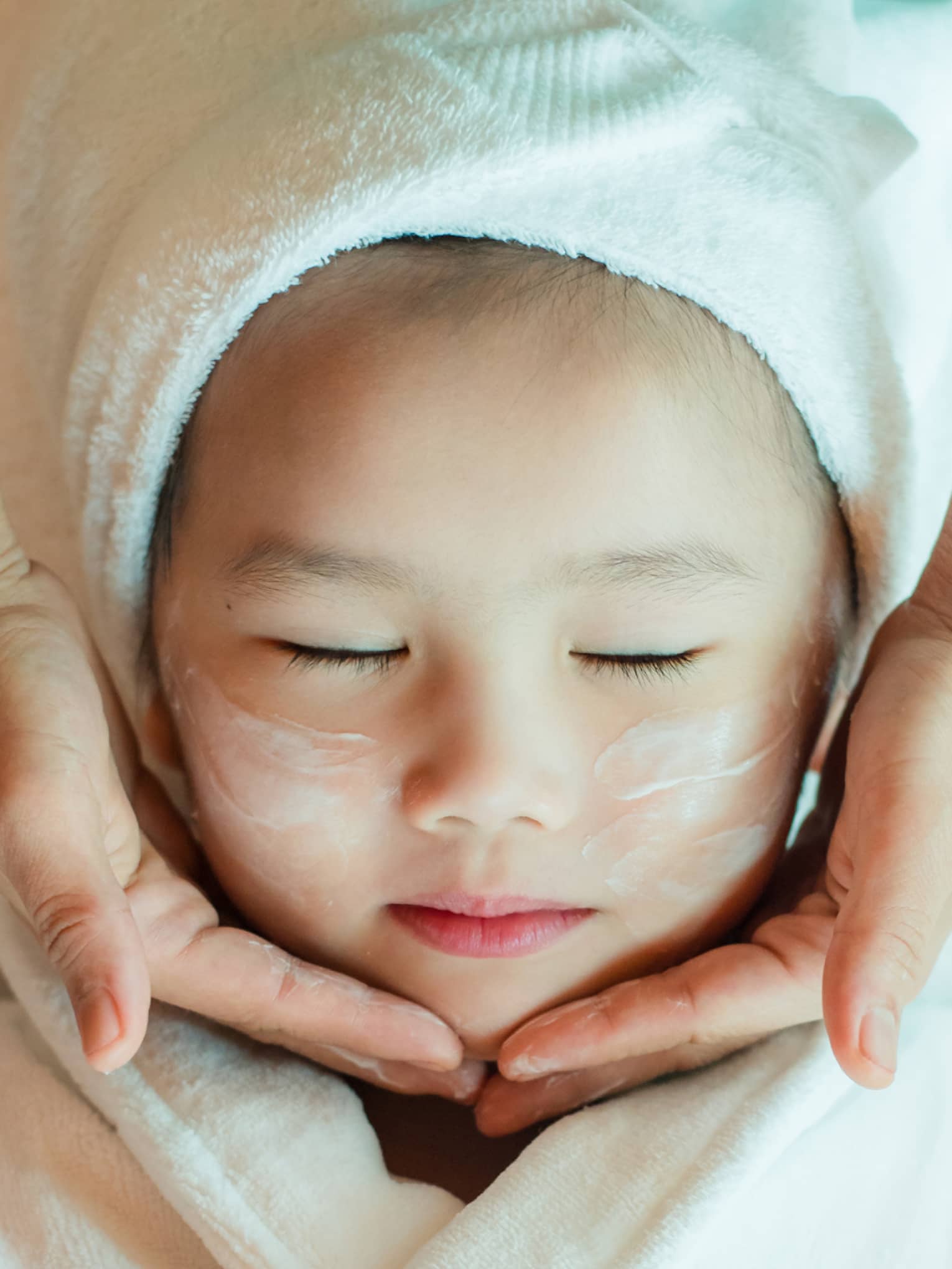 A close-up of a relaxed child wrapped in a towel and robe and enjoying a masseuse massaging a cream into their face.