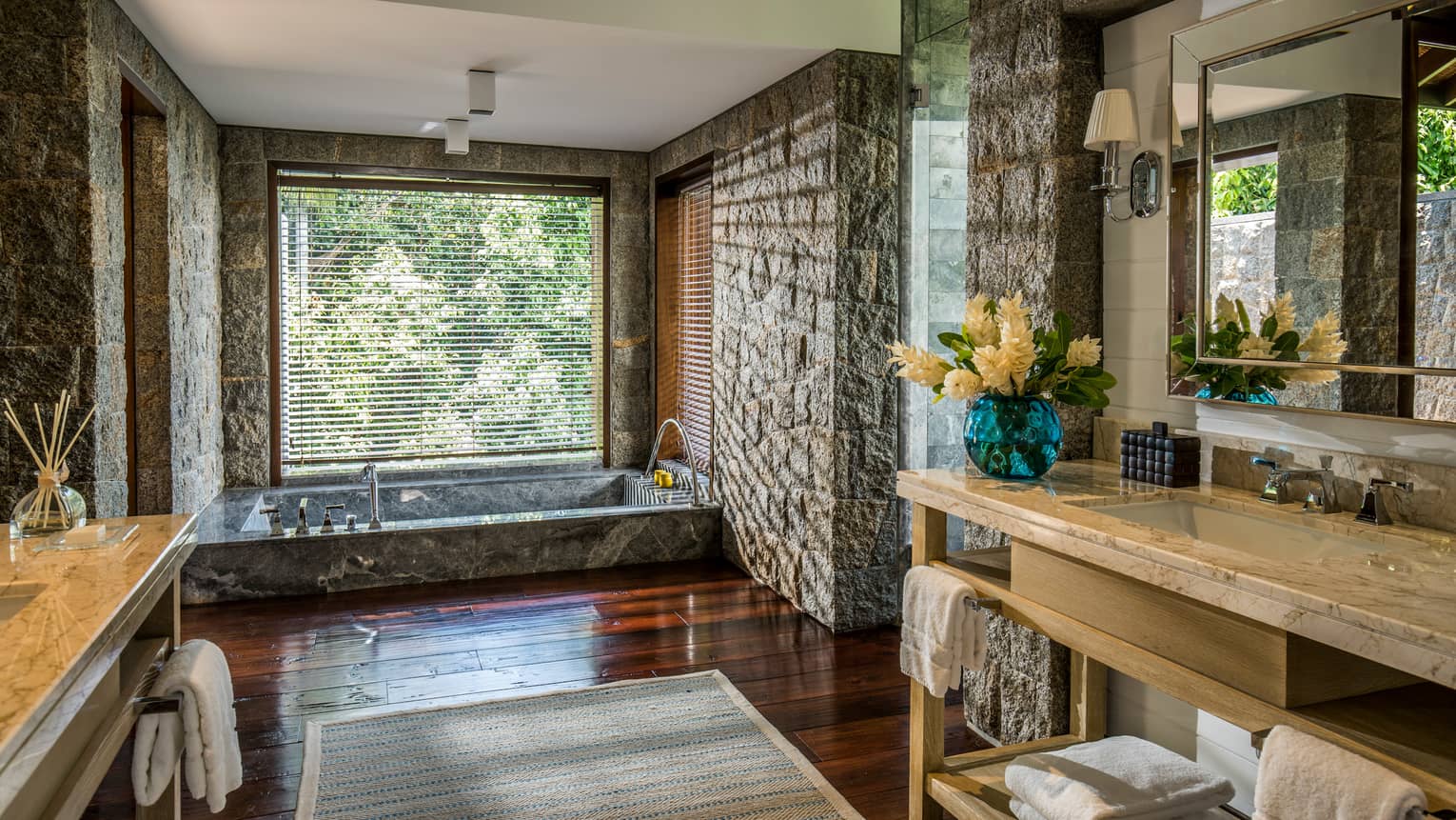 Long vanities in large bathroom with stone walls, sunken marble tub under window