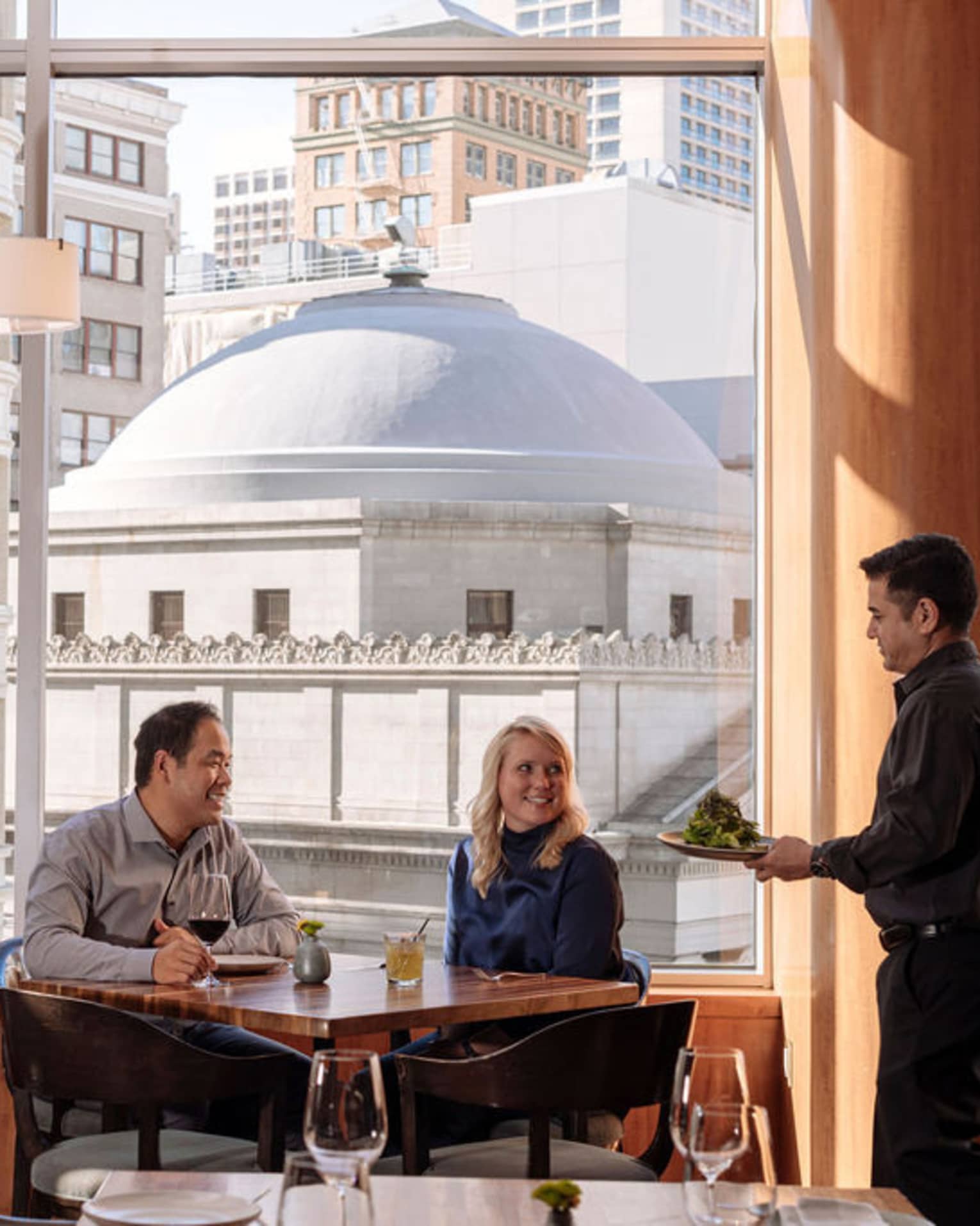 A four seasons staff takes a couple's order as they sit at a window table overlooking the city