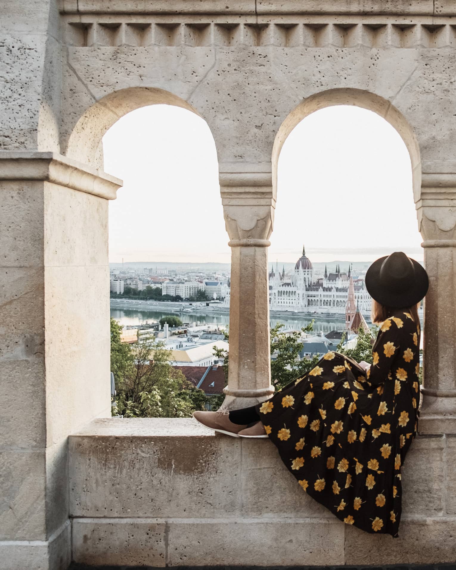 A guest in a floral dress and hat sitting between stone arches, gazing at a distant cityscape and river.