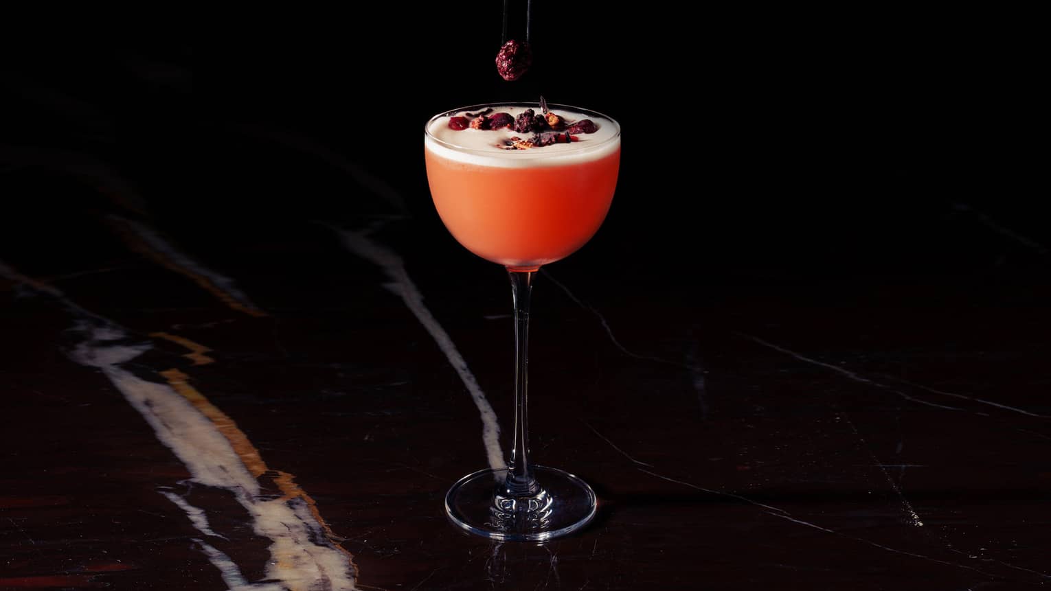 A hand reaches out of the darkness to drop dried cranberries onto a red-colored cocktail in a tall stemmed glass