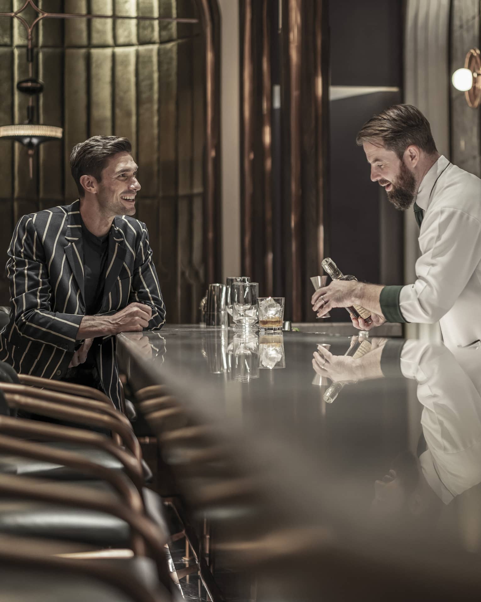 Bartender preparing a drink while a customer sits at the bar, both smiling, in a modern, upscale bar with a reflective countertop and warm lighting.