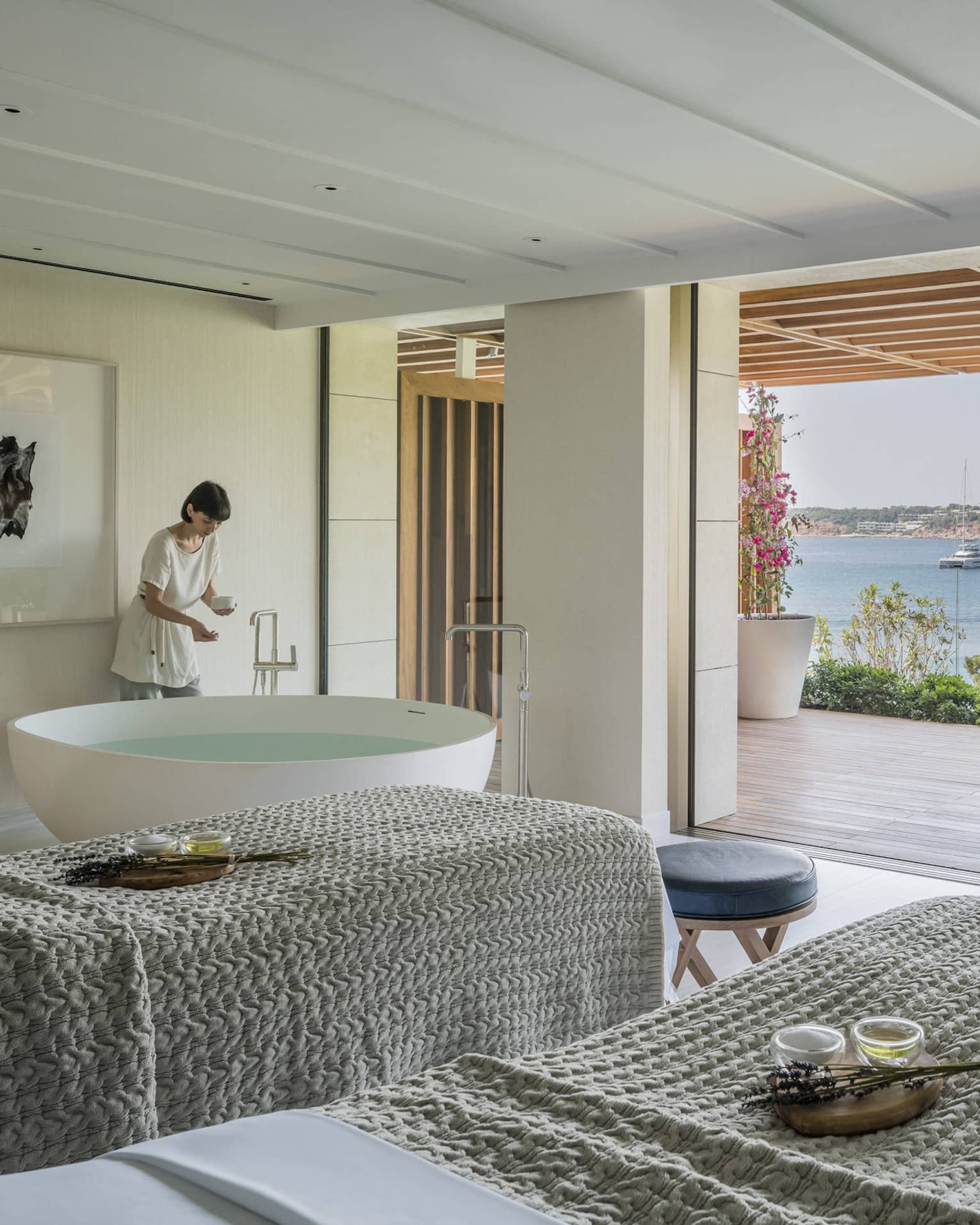 A four seasons staff works in a room with two side by side spa tables draped with gray linens face an open veranda overlooking a body of water in Greece