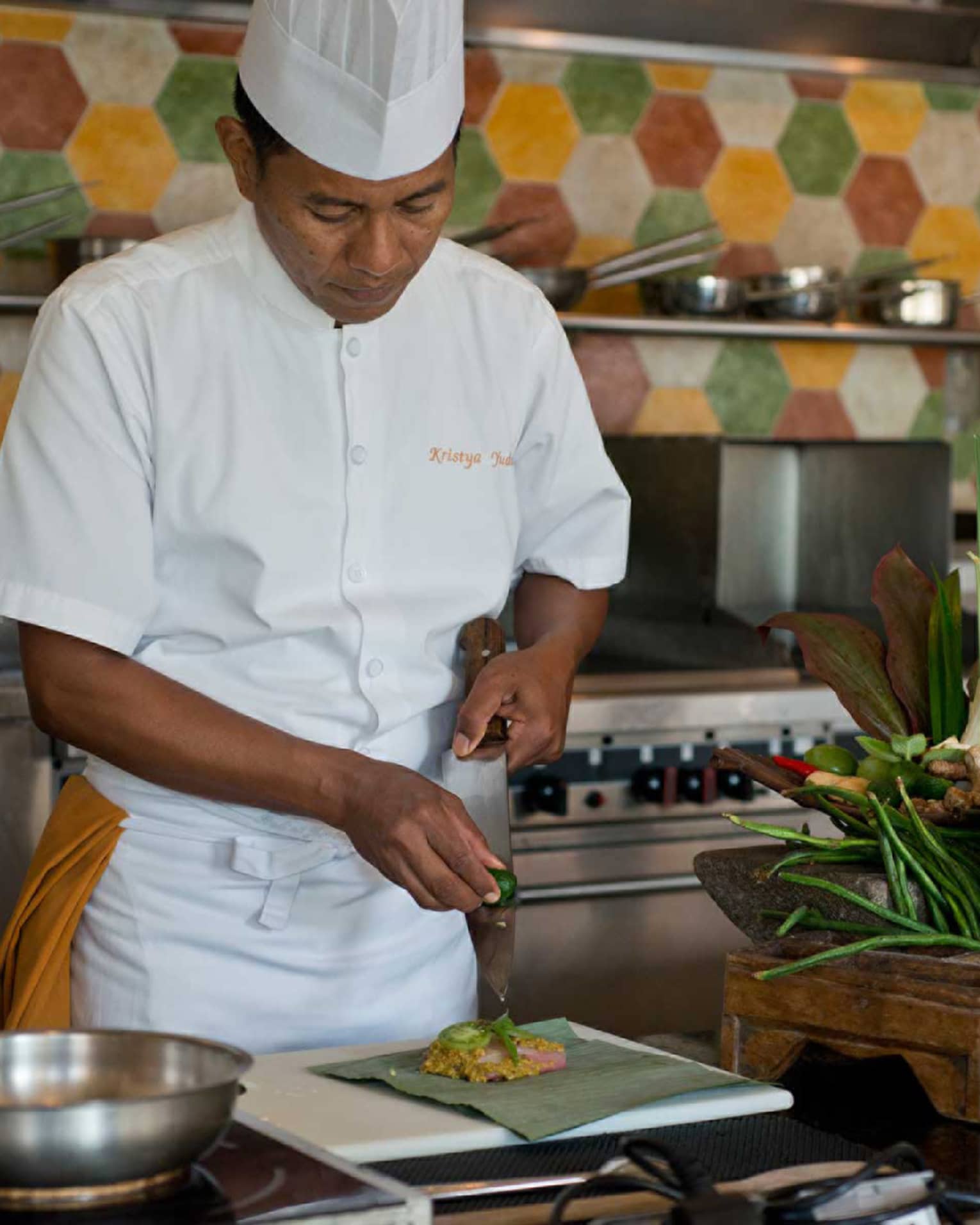 Chef in white uniform squeezes lime on large kitchen knife over dish, next to heads of fresh lettuce and whole peppers