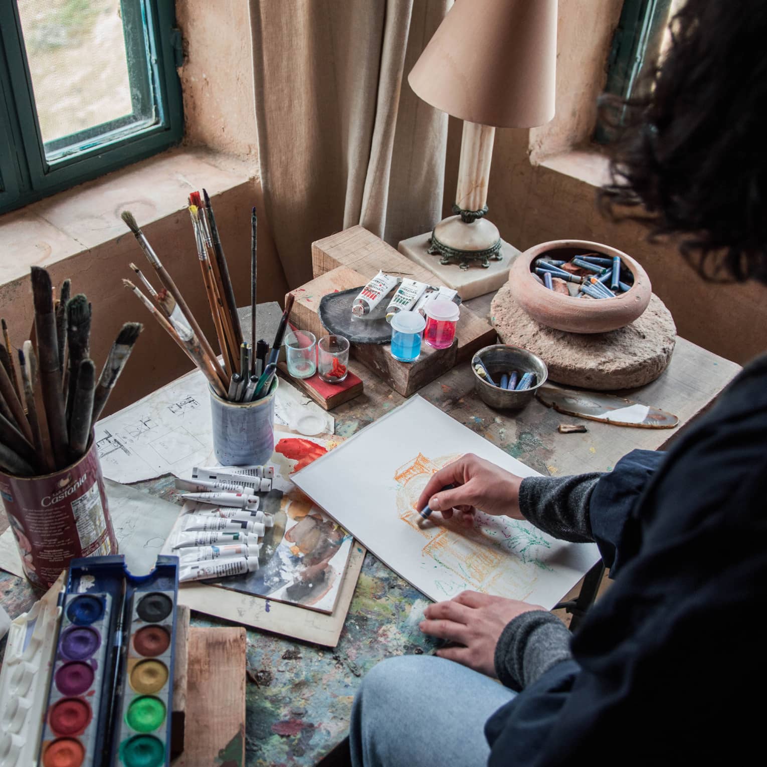 A person with dark curly hair in front of an artists workstation with paintbrushes and acrylic paints.