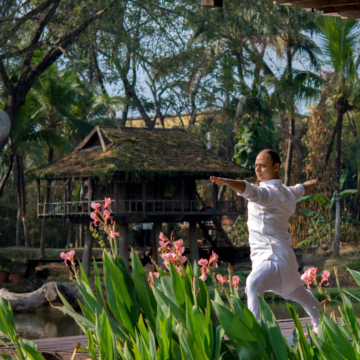 Resident yogi Dheera wearing white in yoga pose with arms outstretched by tall flowers, leaves