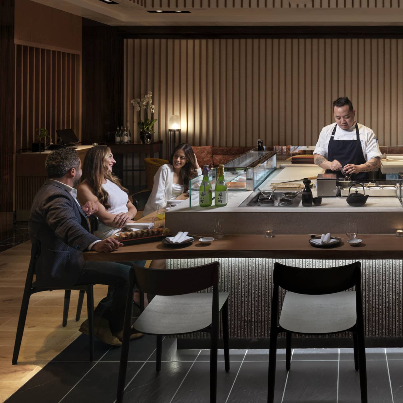Three guests sit at a sushi counter, engrossed in conversation, while two chefs behind the counter carefully prepare food.