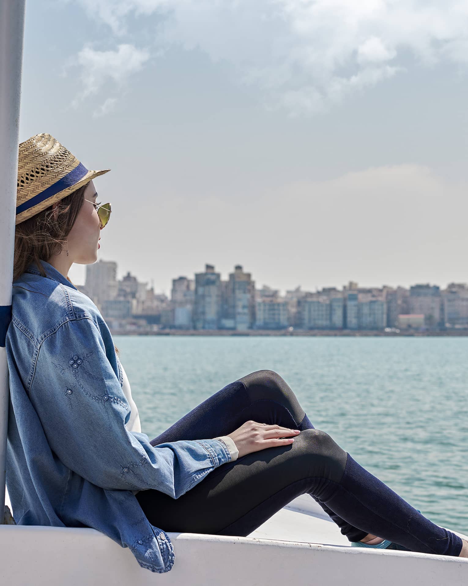 A guest wearing a straw hat sits on the bow of a boat and looks out over calm water toward a distant metropolitan shoreline.