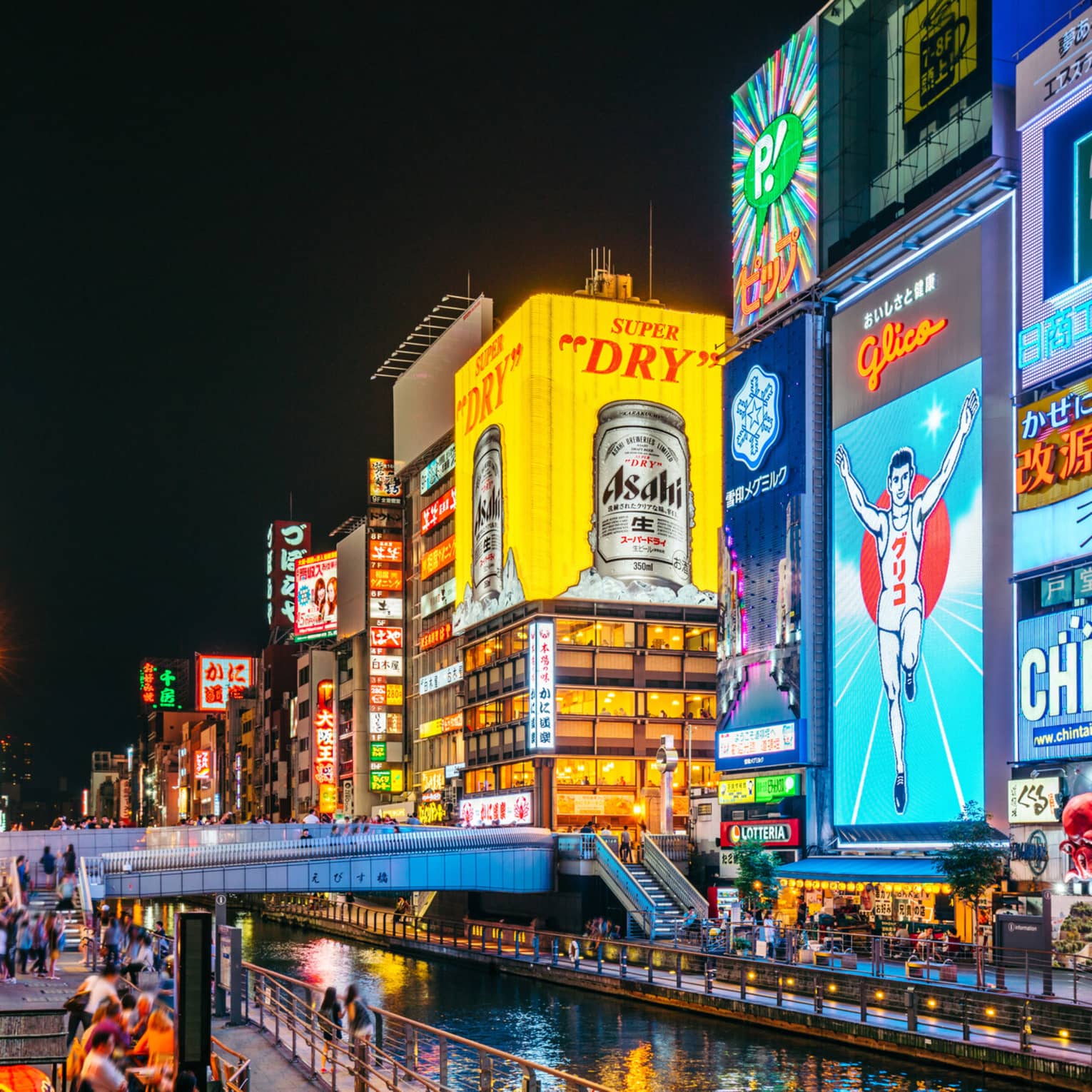 A footbridge crosses a narrow canal flanked by huge neon ads in bright colours on tall buildings in a busy city at night.