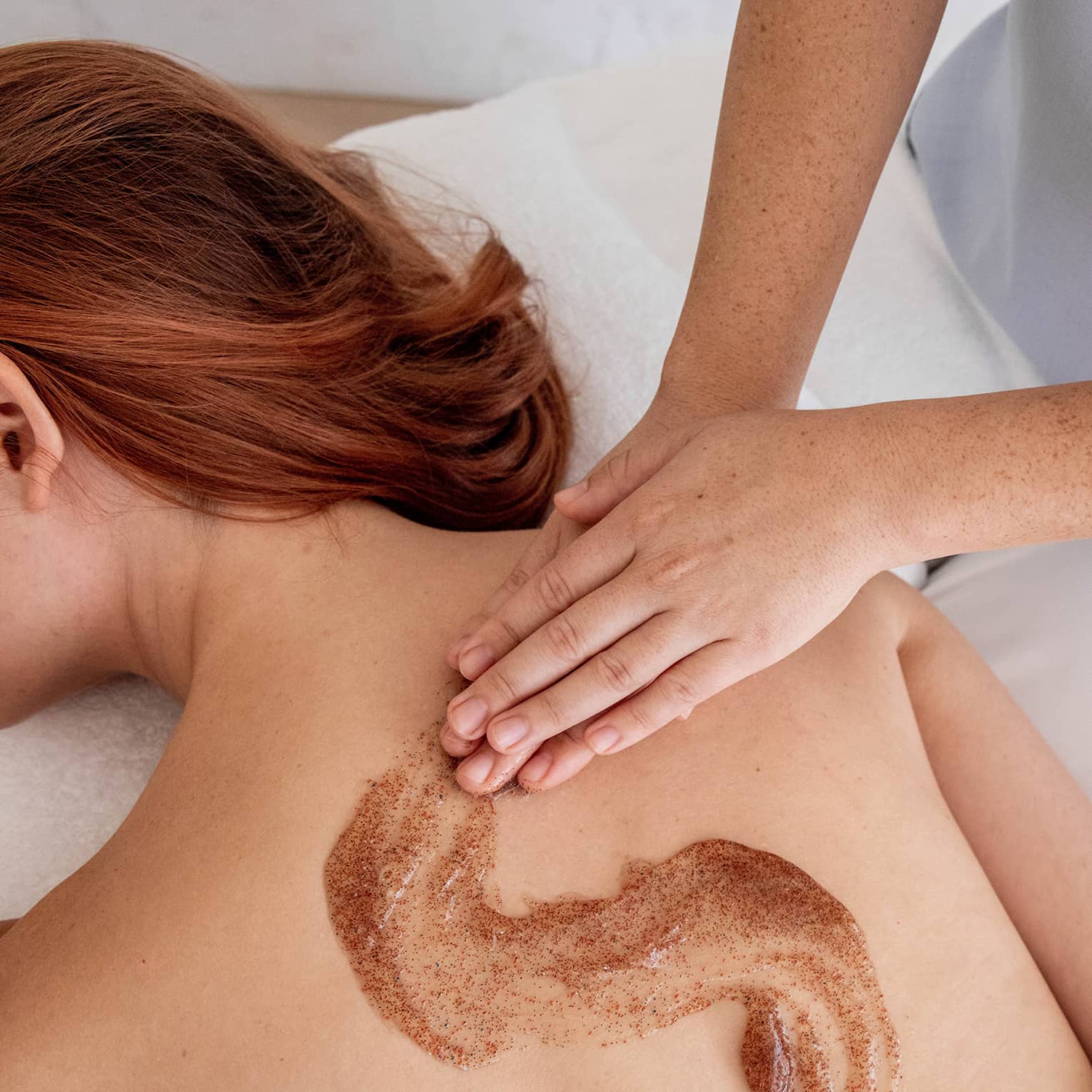 Spa staff massages scrub onto woman's bare back in treatment room