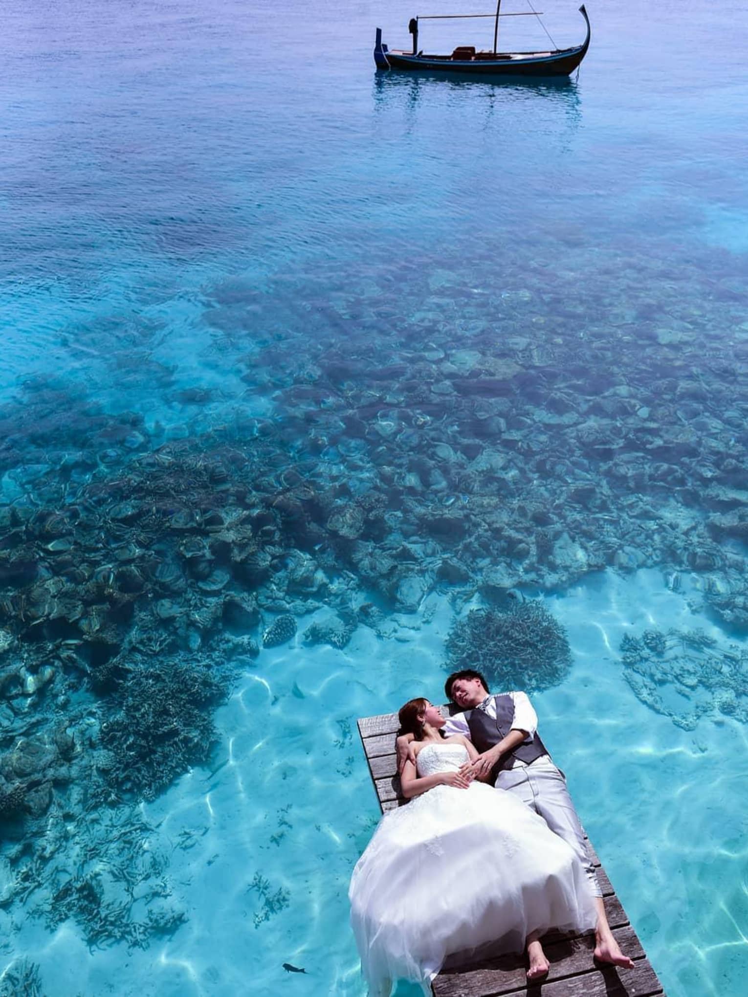Bride and groom lay on dock over blue lagoon, boat in background