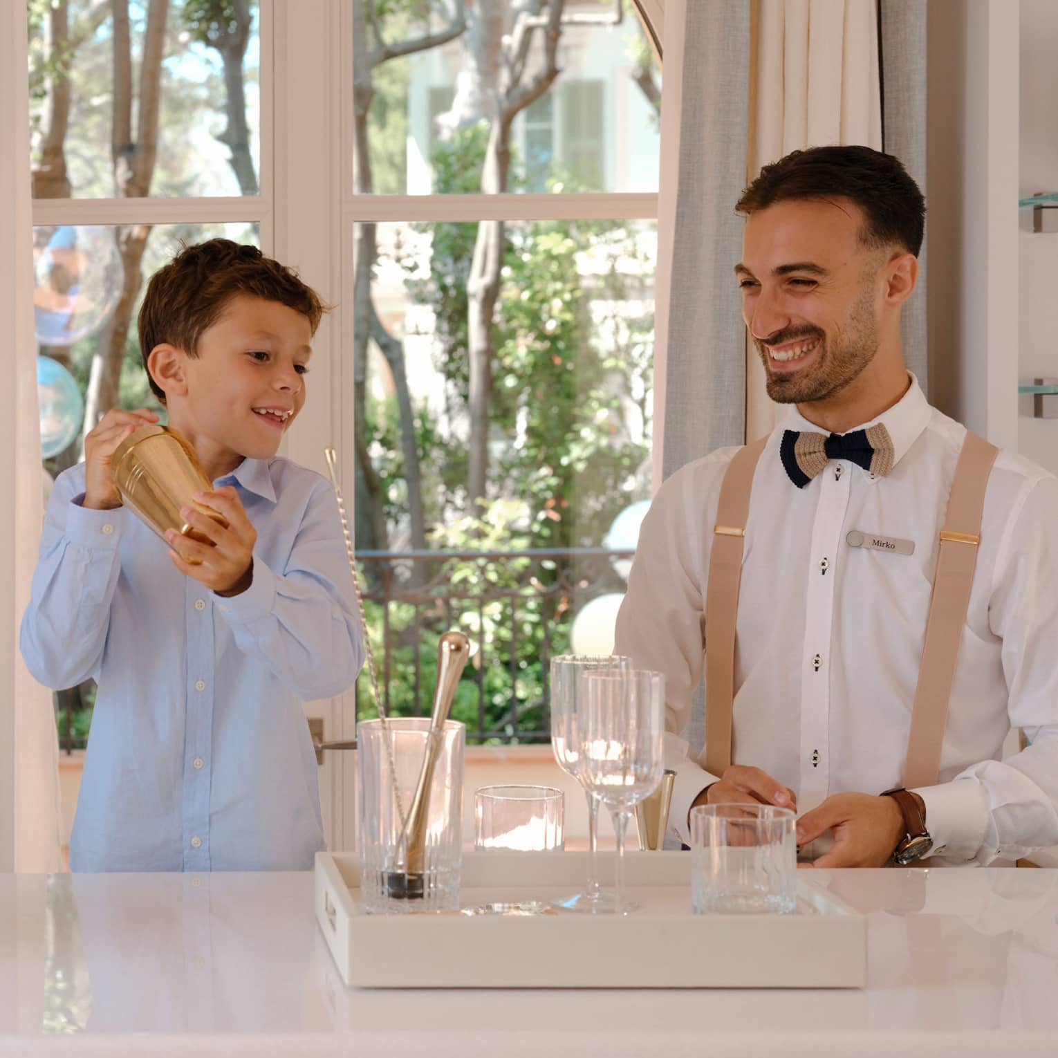 Child holds gold mocktail shaker as smiling bartender looks on