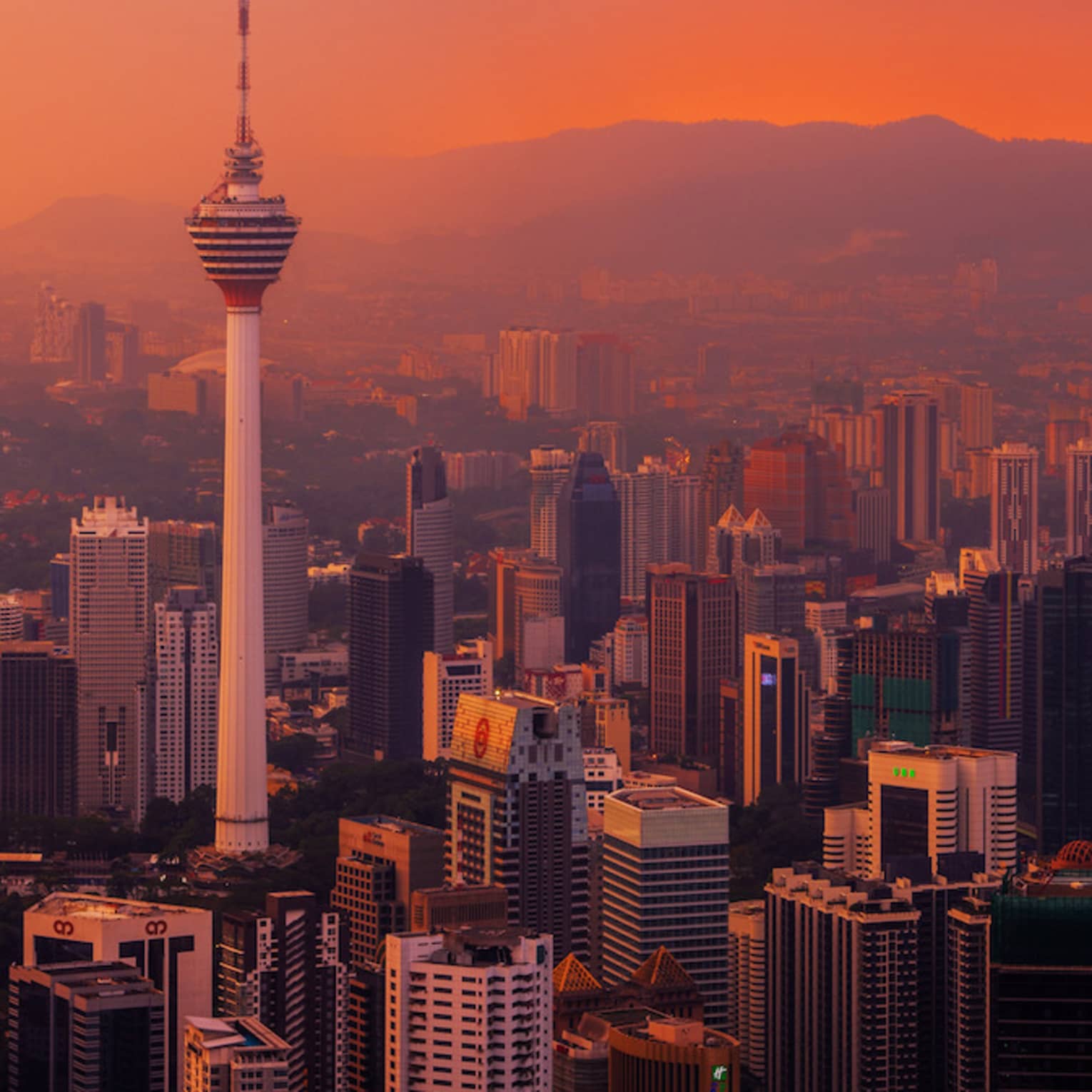 Majestic towers mark the city skyline. Reds and yellows blend in a magnificent sunset as mountains loom in the horizon.