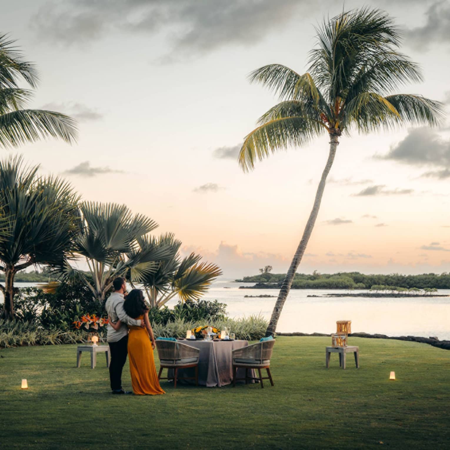 A couple stand on the shore, arms around each other, gazing toward the ocean; set table with two chairs beside swaying palms.
