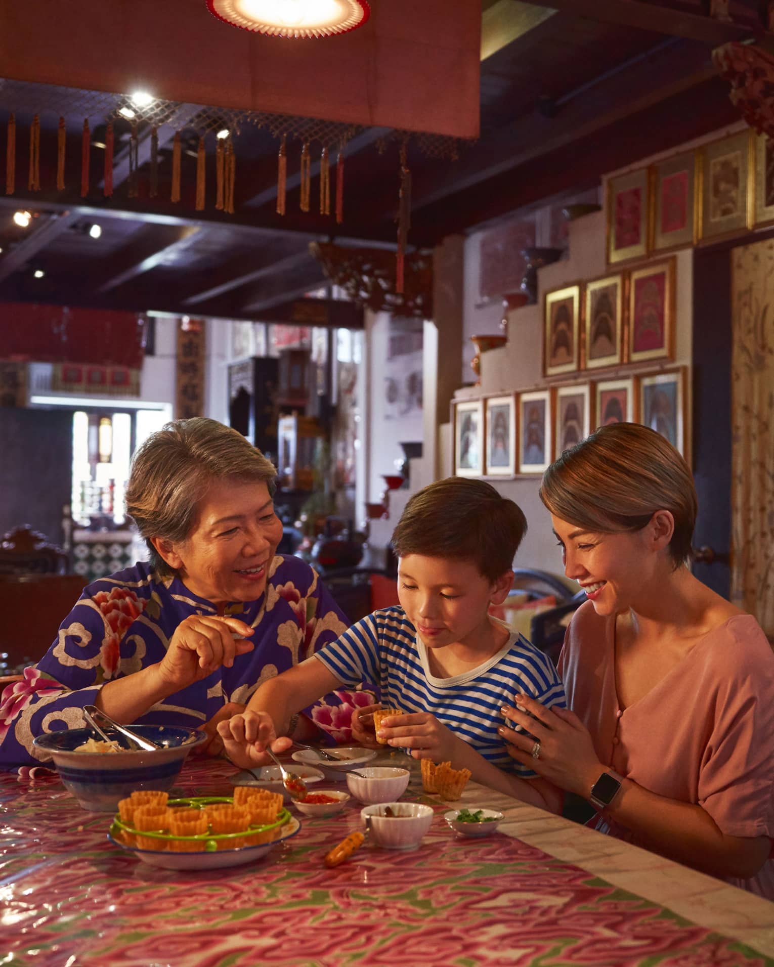 A family sits in a richly decorated, dimly-lit room at a table with various bowls as a child scoops jam into a pastry shell.