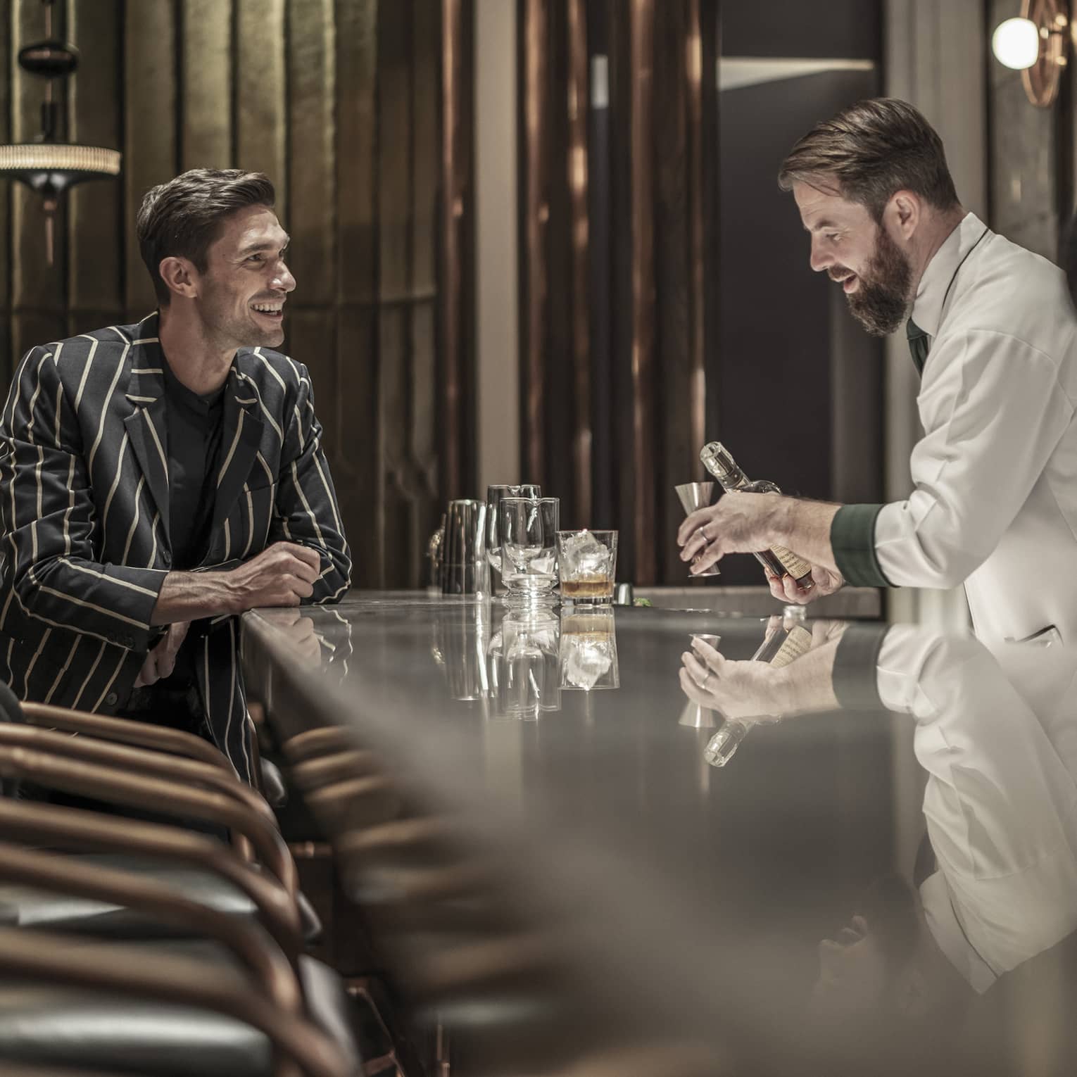 Bartender preparing a drink while a customer sits at the bar, both smiling, in a modern, upscale bar with a reflective countertop and warm lighting.