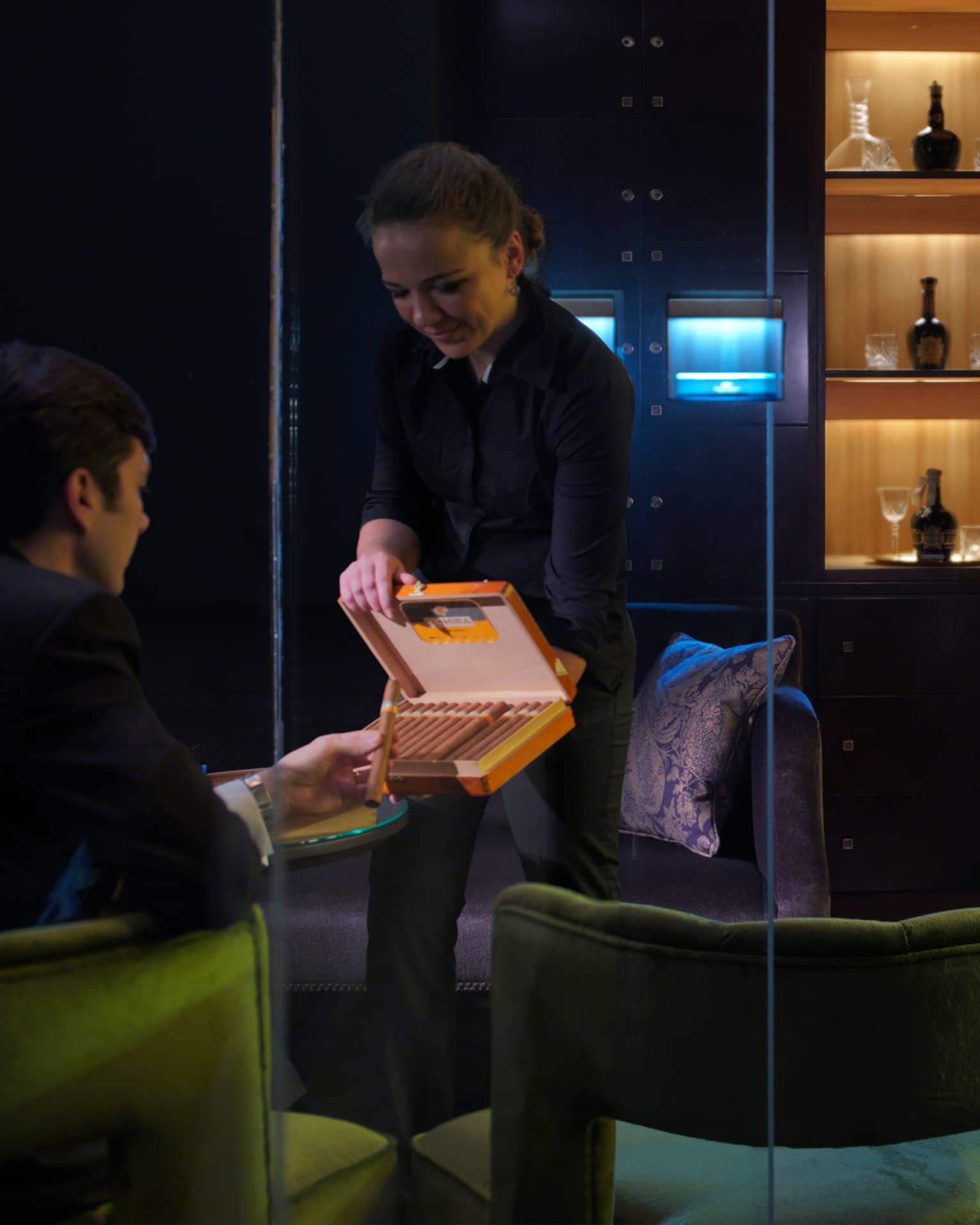 Woman holds open box of cigars, displays them to man sitting in dark lounge