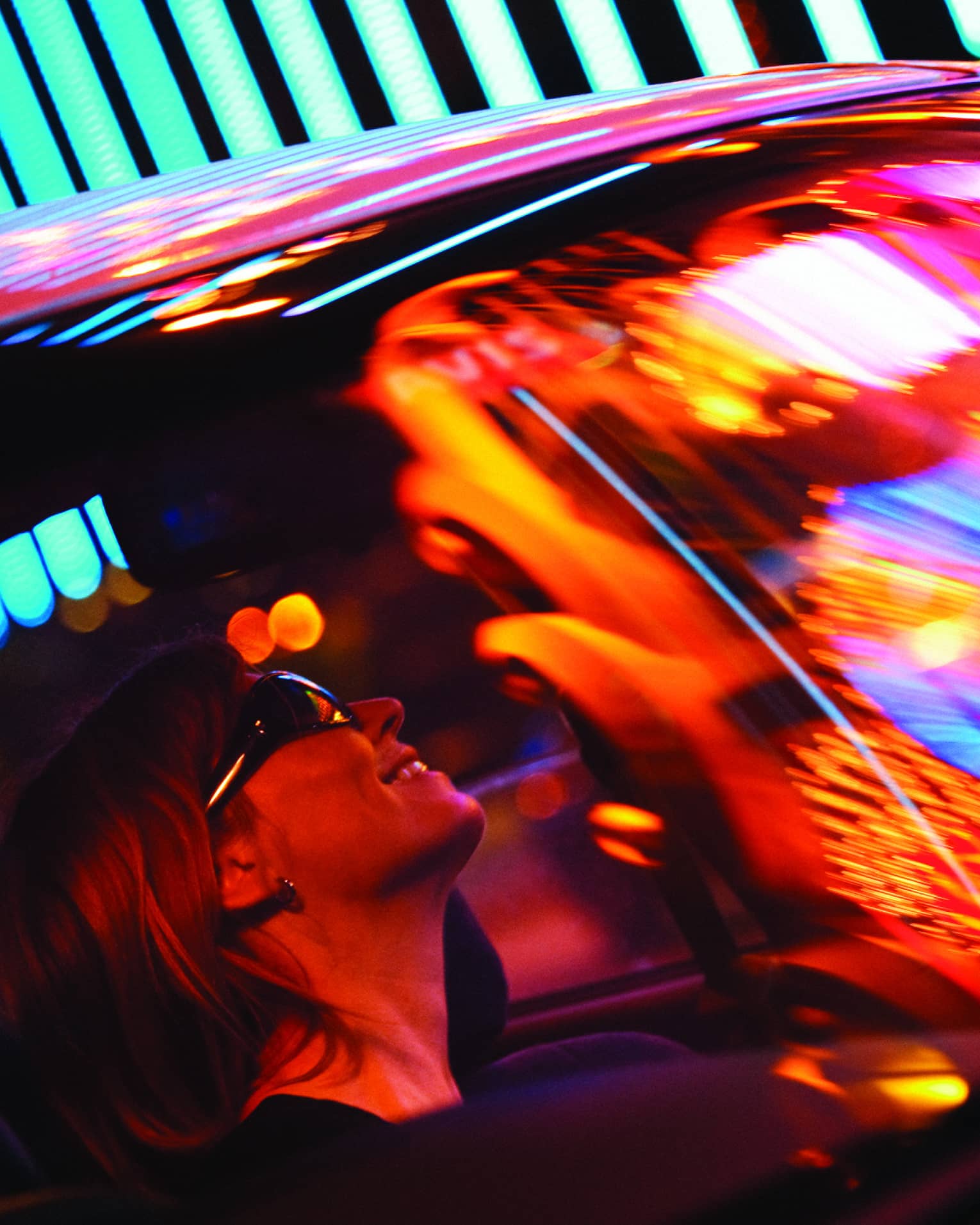 View of woman wearing sunglasses through car windshield reflecting city lights at night