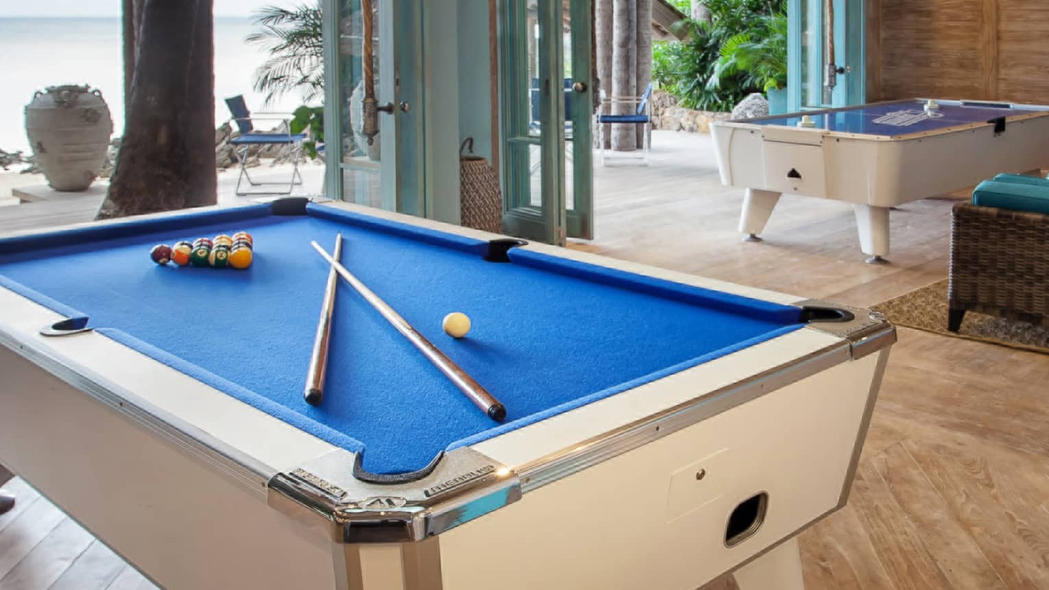 Blue billiards table with pool balls, sticks by seating area, open wall to patio