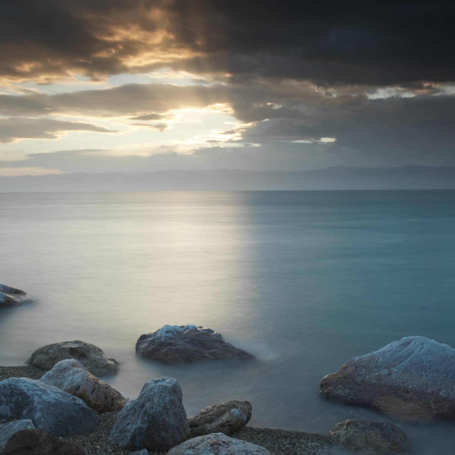 By a stony shoreline, rocks jut out of a dark lake beneath a beam of sunlight filtering through a hole in dark grey clouds.