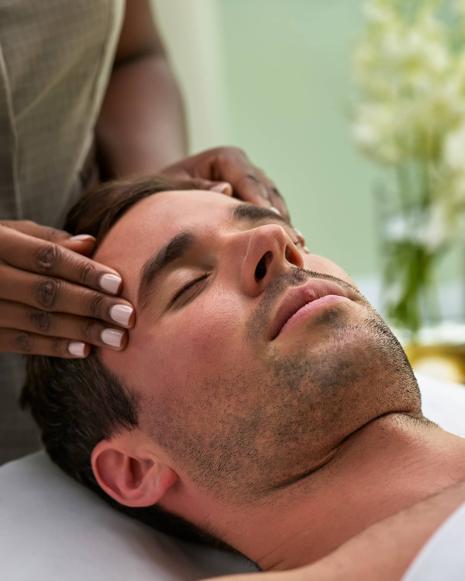 Man lies on massage table with eyes closed as hands massage his temples, forehead