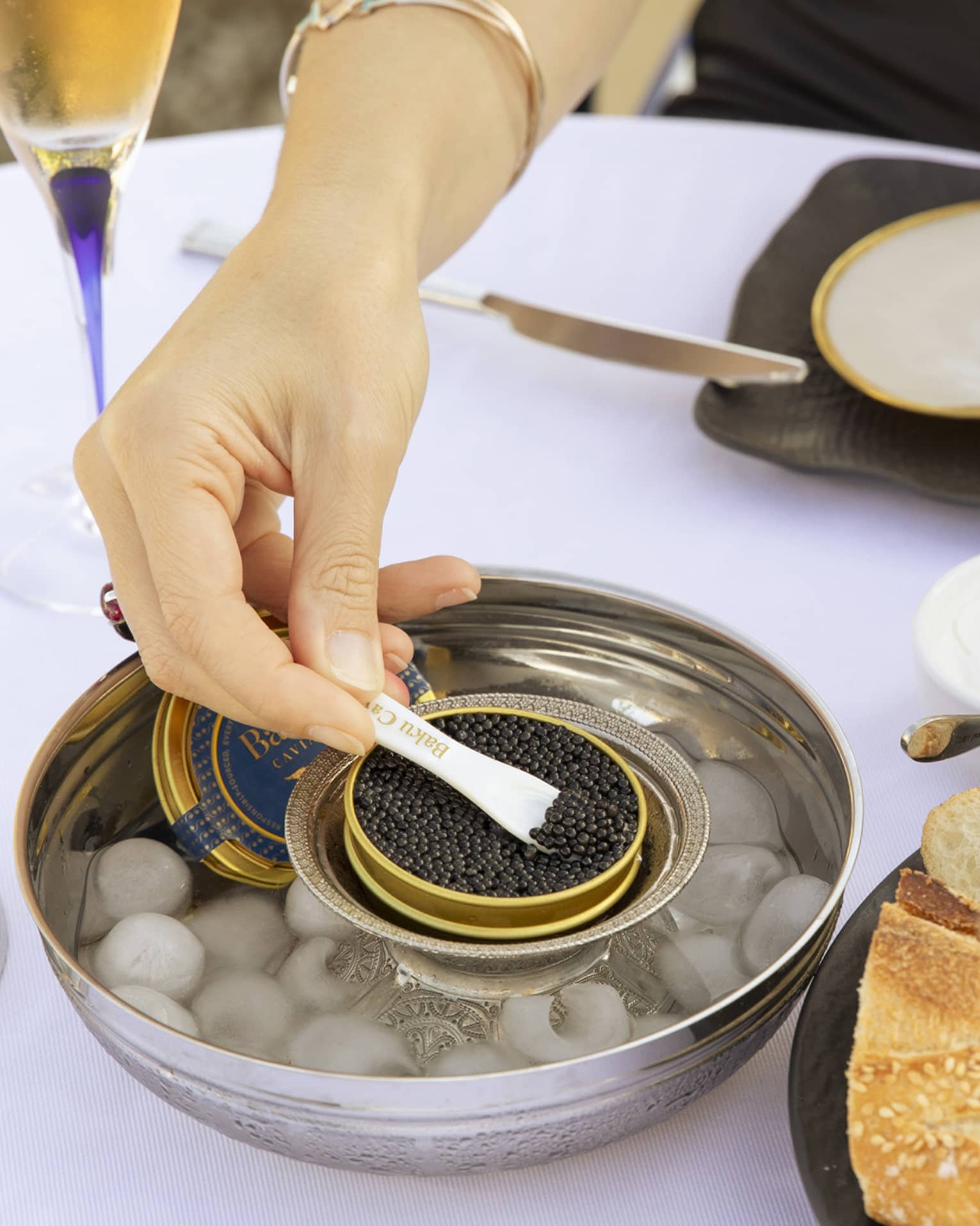 At a table set with champagne flutes, a diner holds buttered bread in one hand and spoons black caviar with the other.