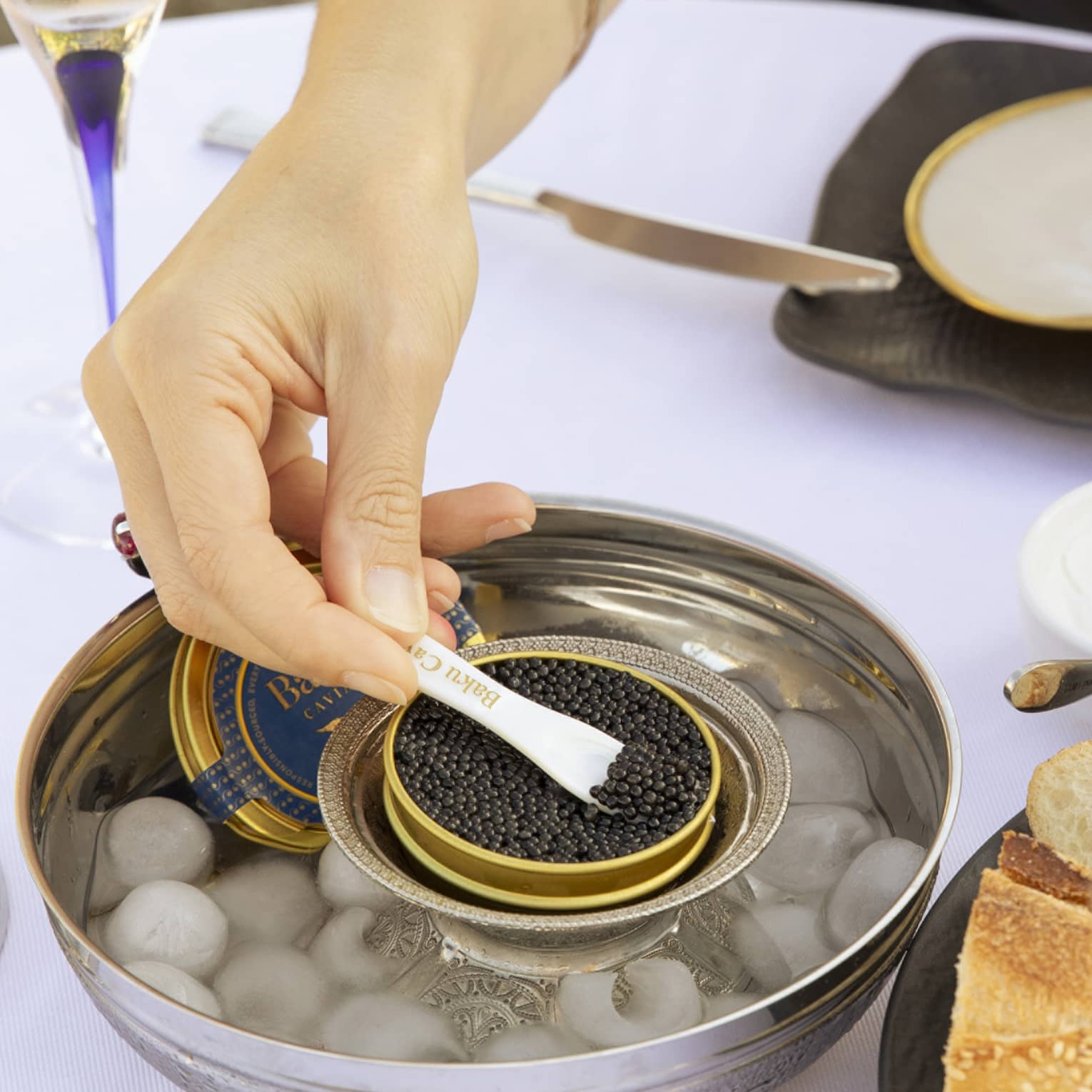 At a table set with champagne flutes, a diner holds buttered bread in one hand and spoons black caviar with the other.