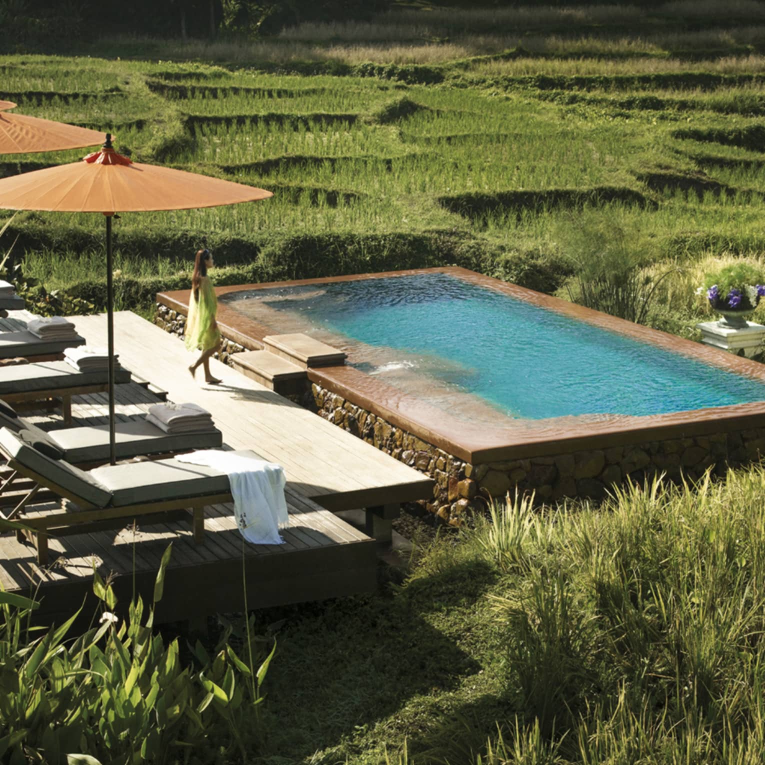 Woman in flowing dress walks past three patio umbrellas to infinity pool in rice plantation