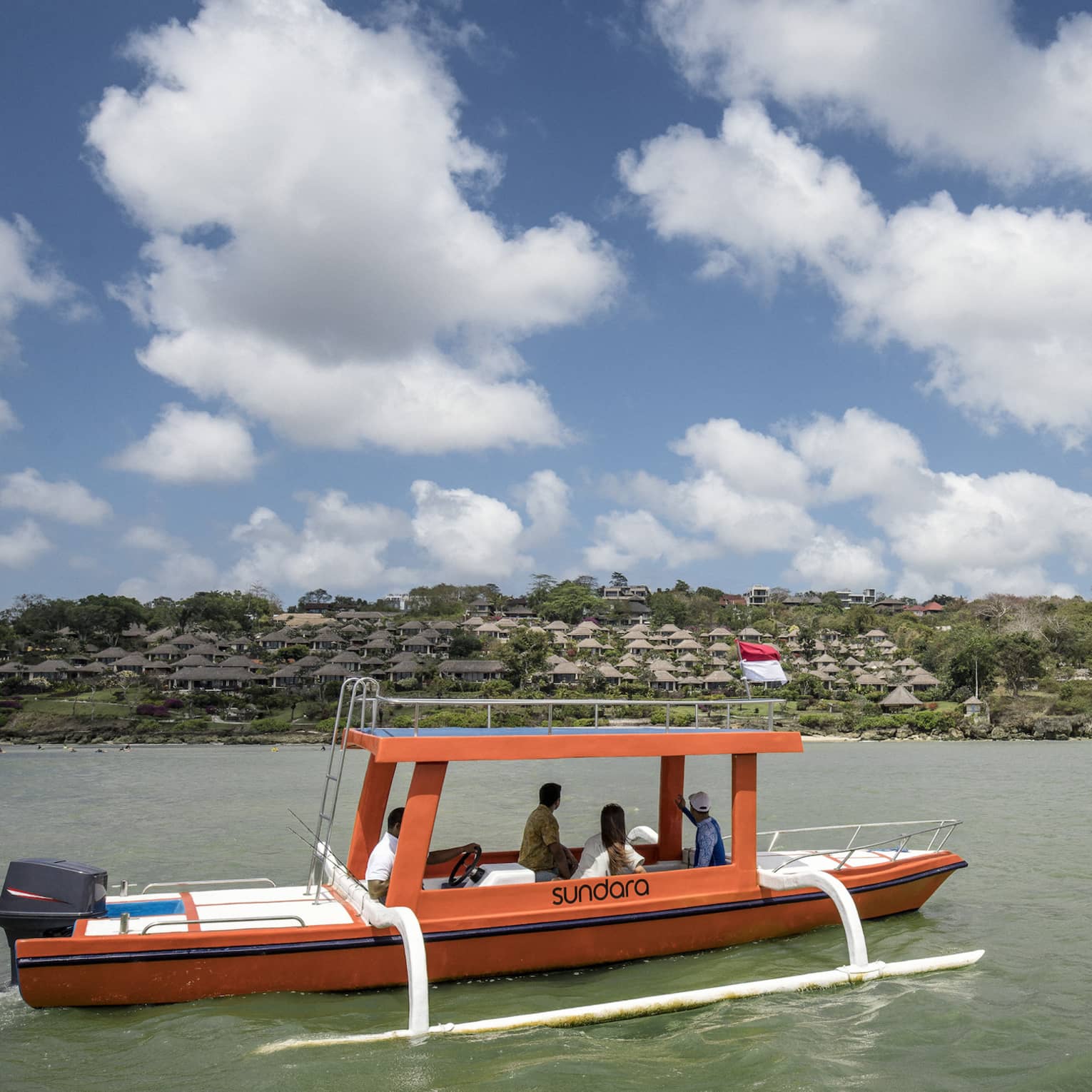 An outrigger canoe glides along calm, green water as four passengers look toward a distant shore dotted with trees and huts.