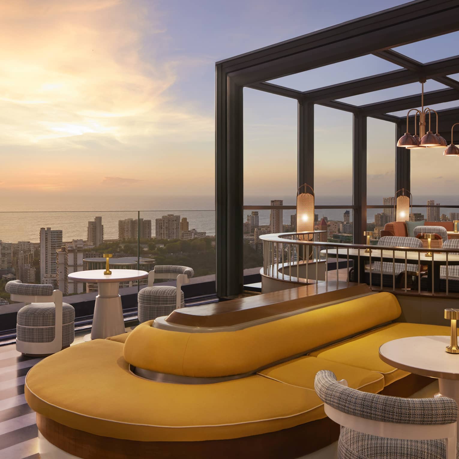 A modern rooftop patio with a mix of booth bank seating and tables for two, panoramic cityscape view and dusky ocean beyond.