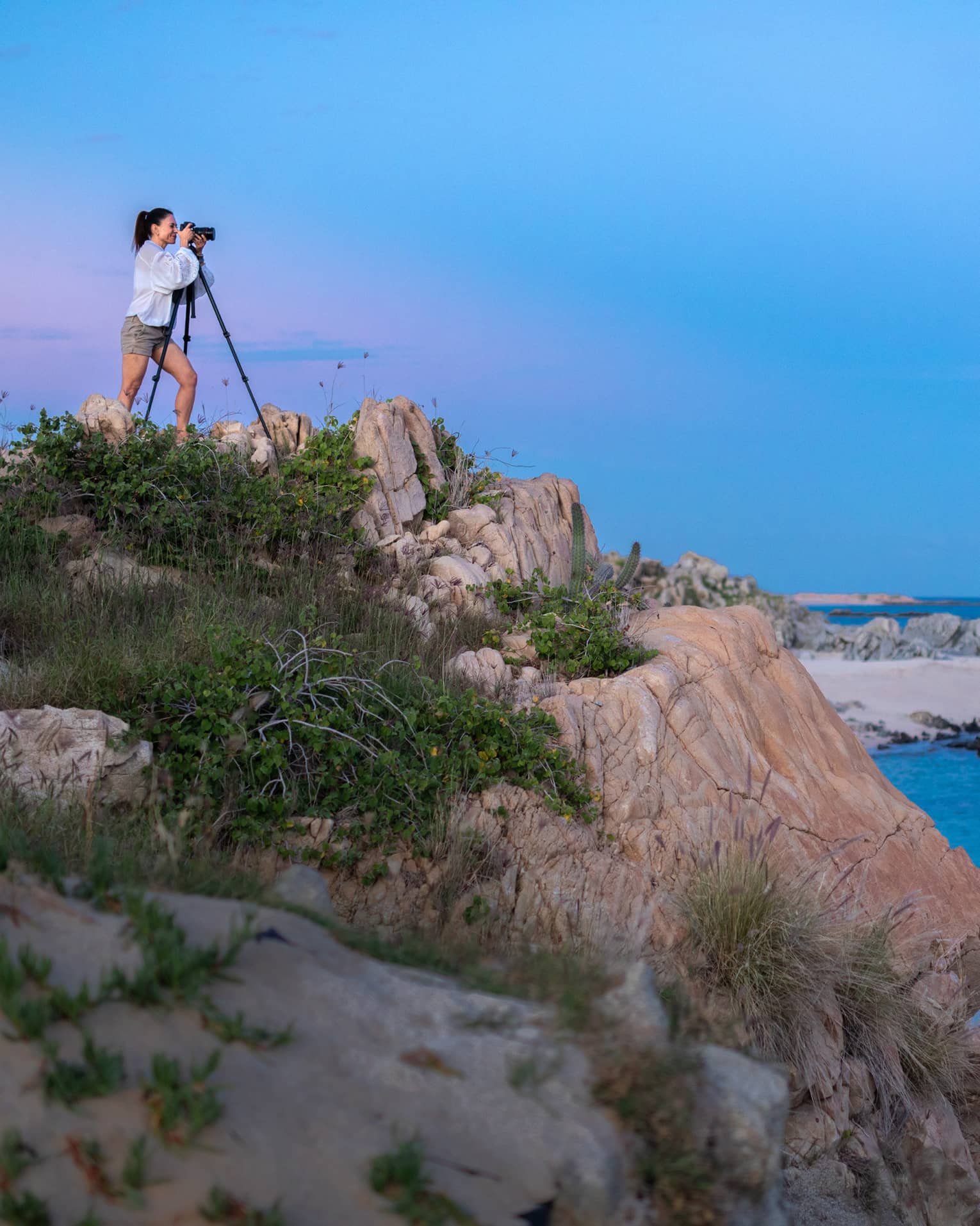 A photographer squints into a camera set on a tripod on a grassy rock outcrop overlooking calm, dark water under a full moon.