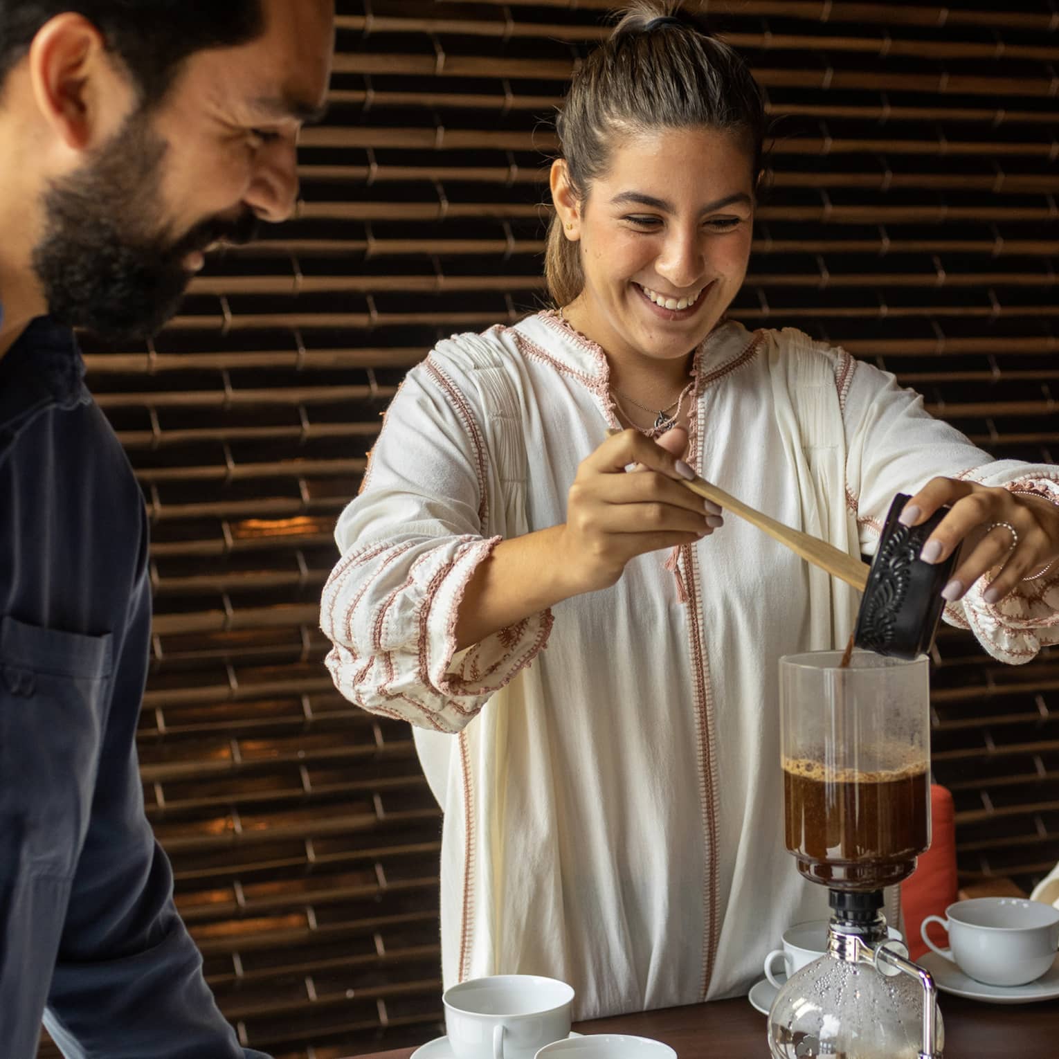 At a table set with white cups and saucers, two smiling guests watch as another pours coffee into a siphon coffee maker.
