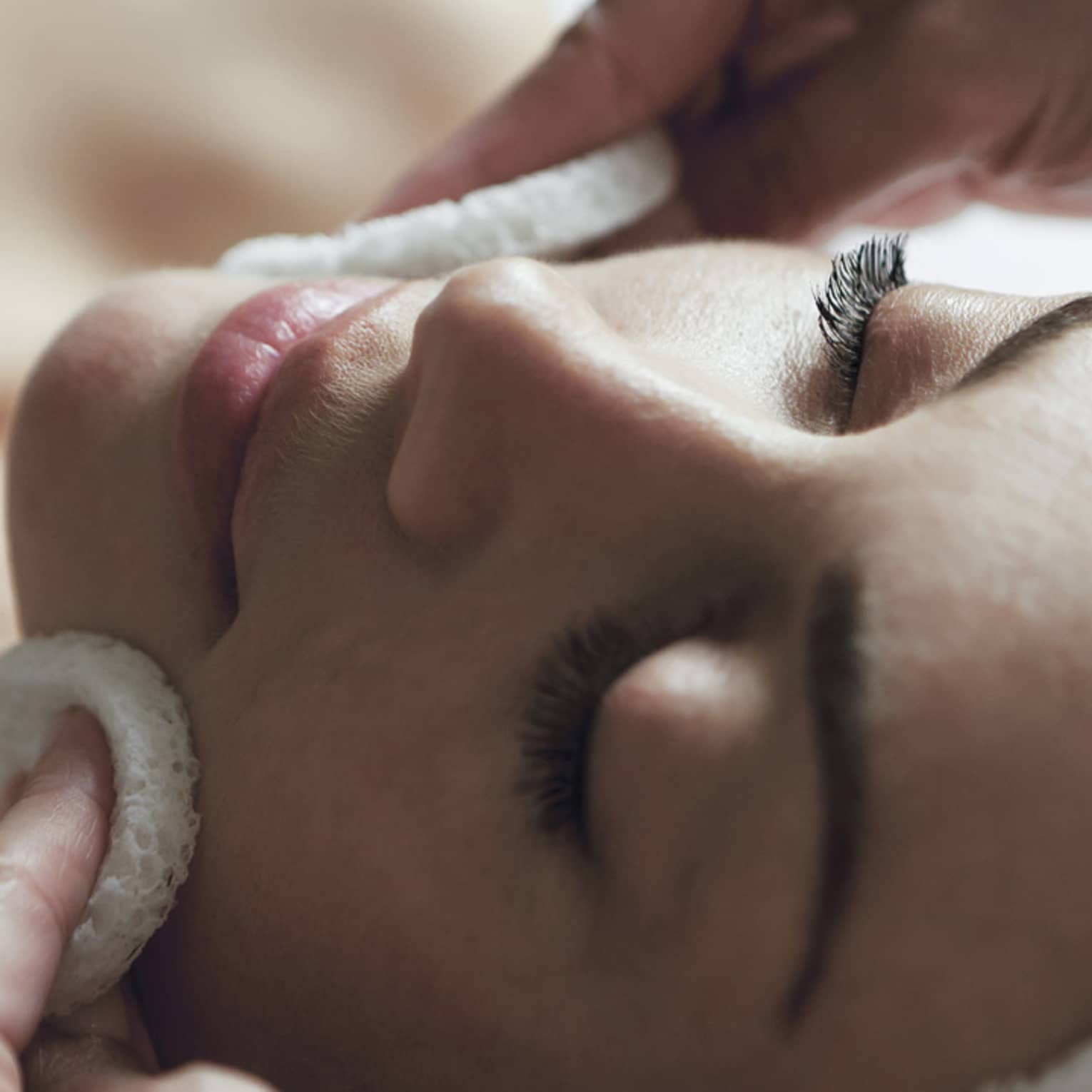 Woman with eyes closed, hair wrapped in towel, as spa attendant rubs cheeks with white pads