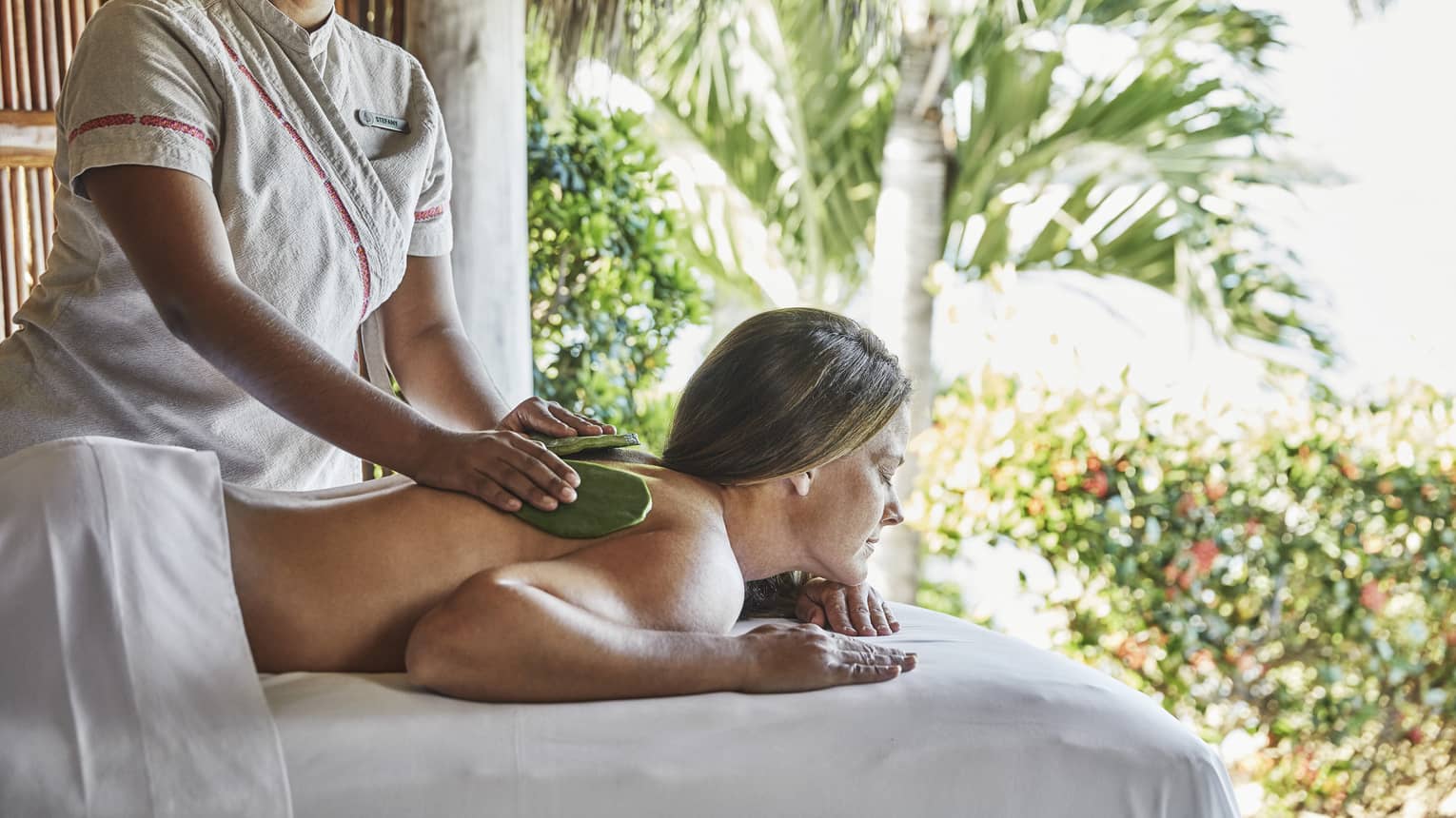 Therapist performs Lomi Mohala massage on woman on massage table, outdoors under thatched hut