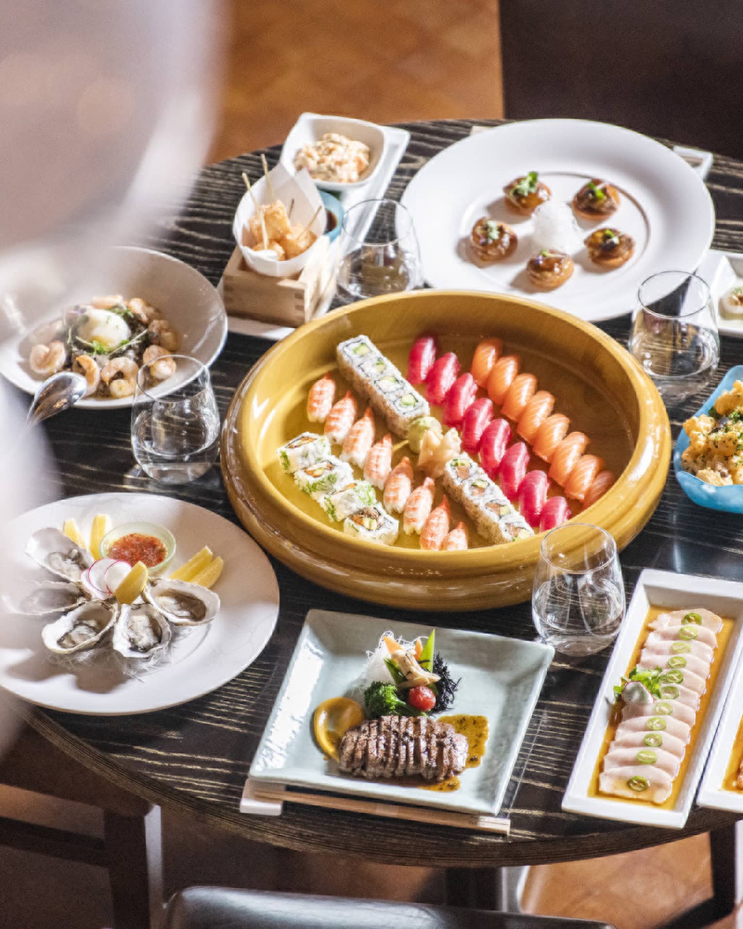 Small round table filled to the brim with a variety of square and round dishes filled with food, all surrounding a round gold platter with five rows of sushi in the center