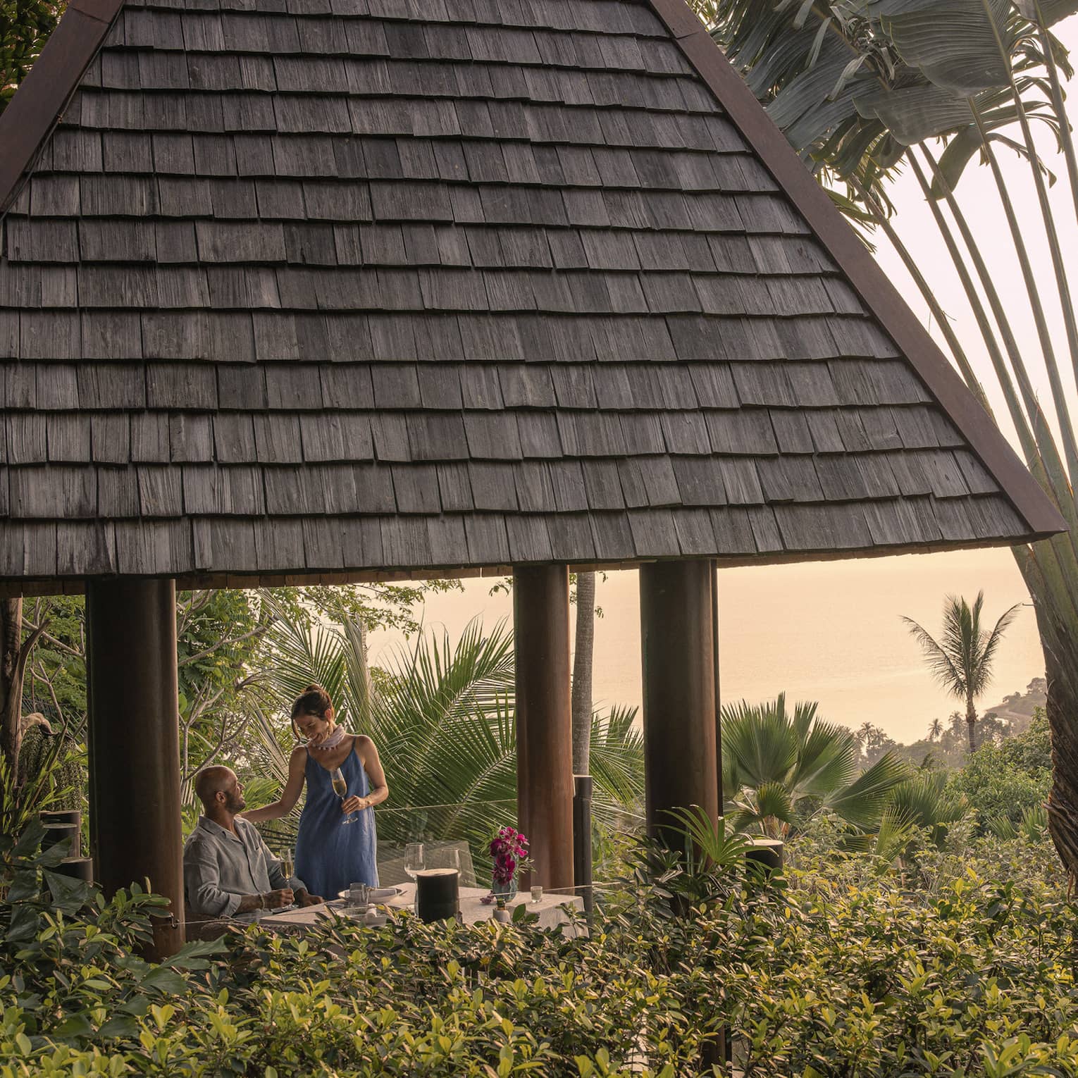 A couple with champagne in a tall hilltop pavilion with a dark, straw roof, set amidst lush forest and a giant fan palm tree.