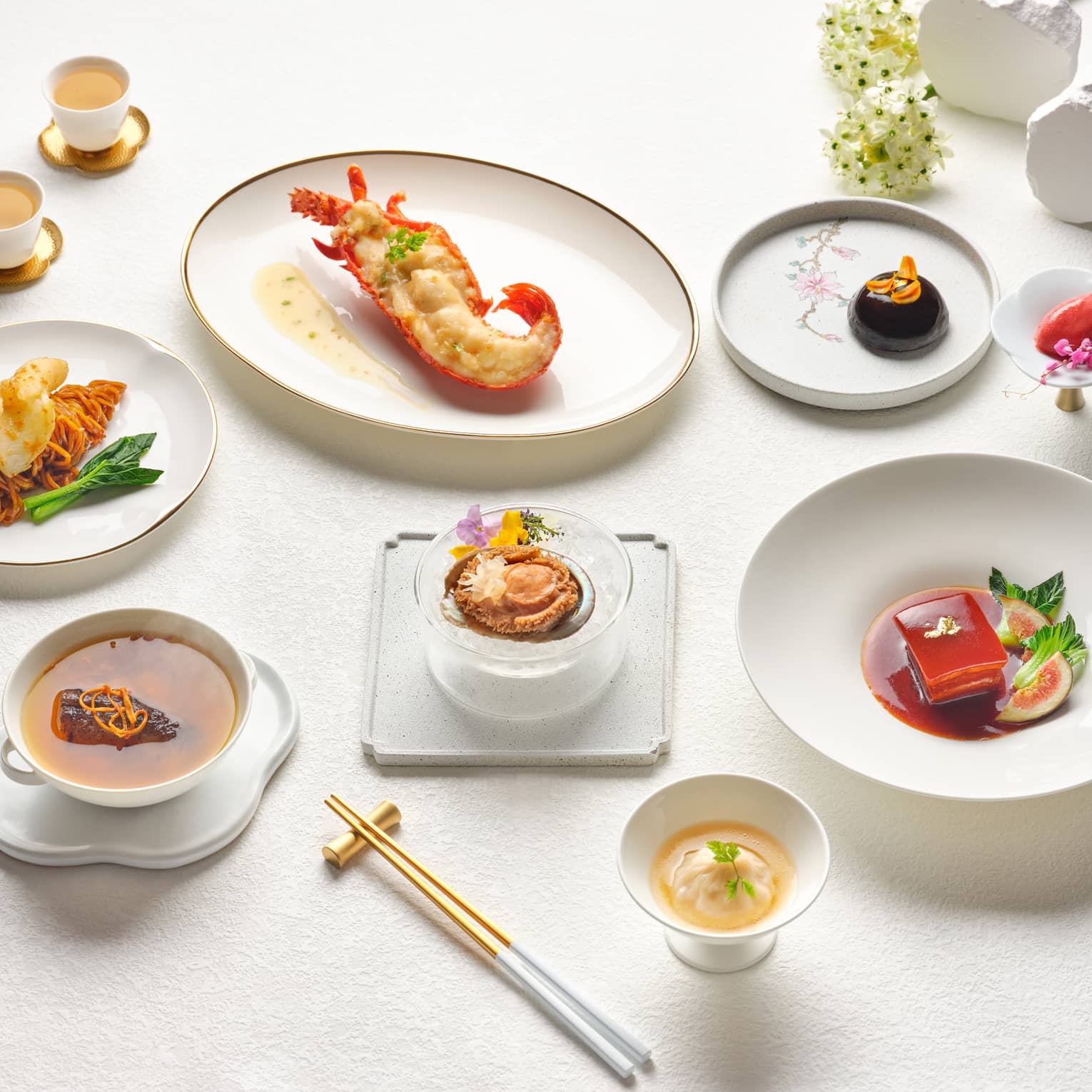 A beautifully arranged gourmet Chinese meal featuring lobster, braised meat, dumplings and dessert, presented on elegant white tableware with teacups and gold chopsticks.