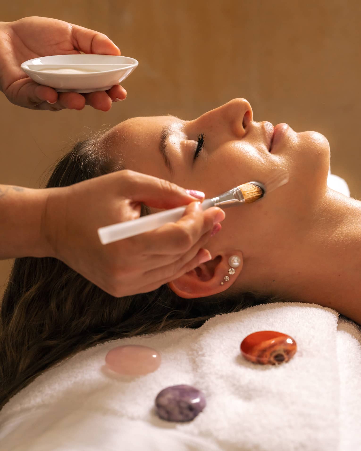 Woman lies covered by white blanket from shoulders down as someone gently applies a cream on their face with a brush, with three gemstones places next to the client's head