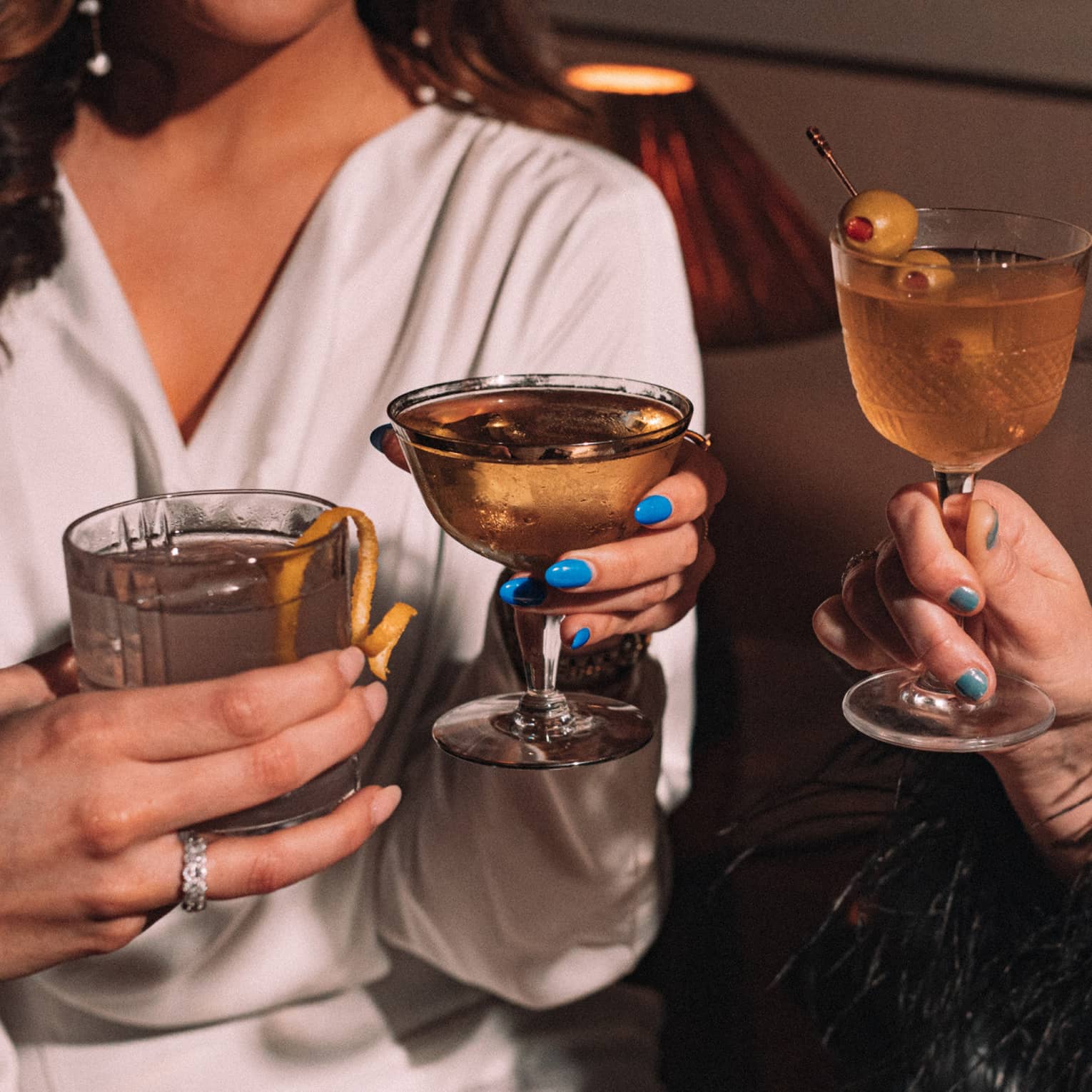 Three smiling guests share a toast with stemmed glasses and tumblers of cocktails with olive and lemon garnish.