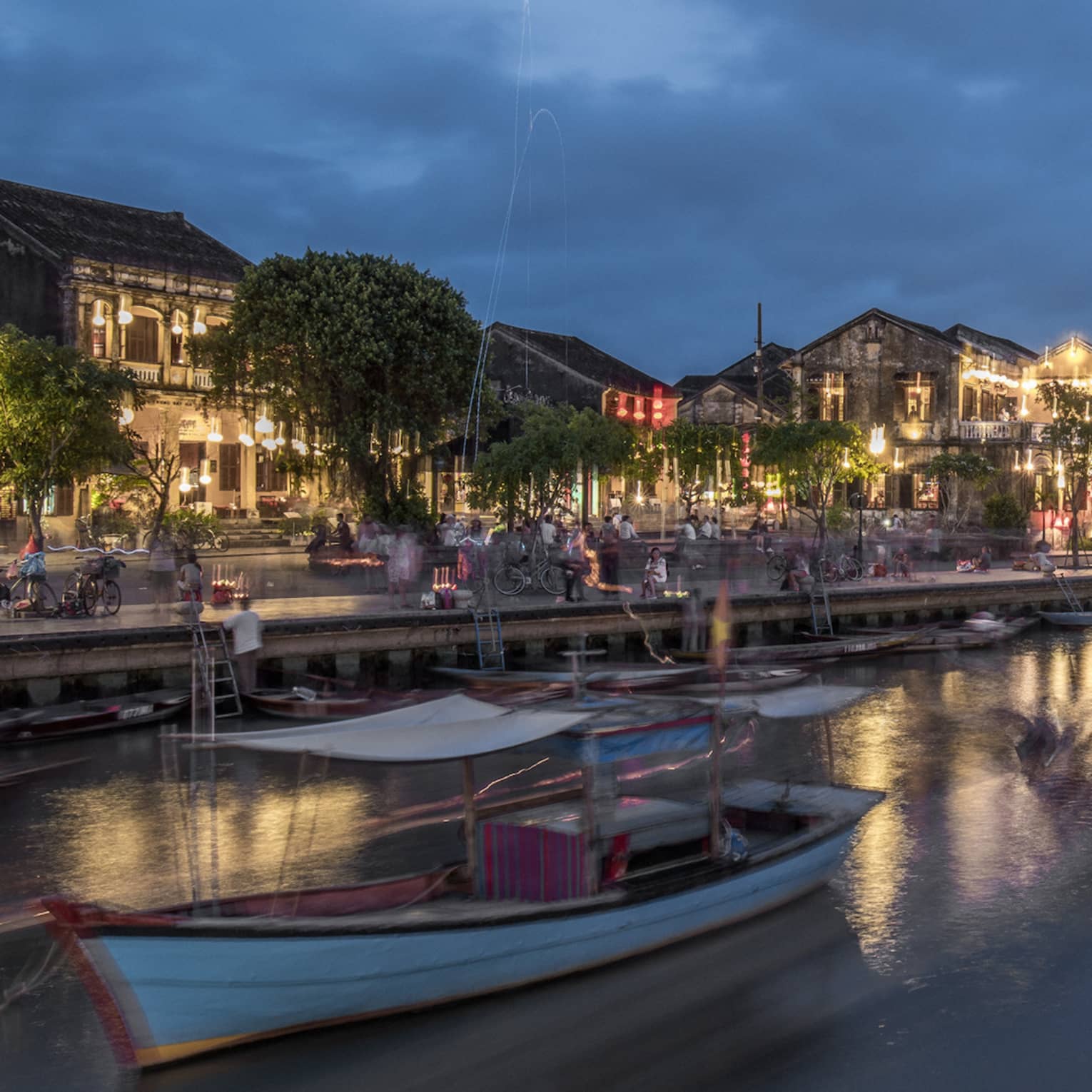 Evening view of a bustling riverside town with boats moving on the water, illuminated by streetlights and buildings along the waterfront, under a deep blue sky.