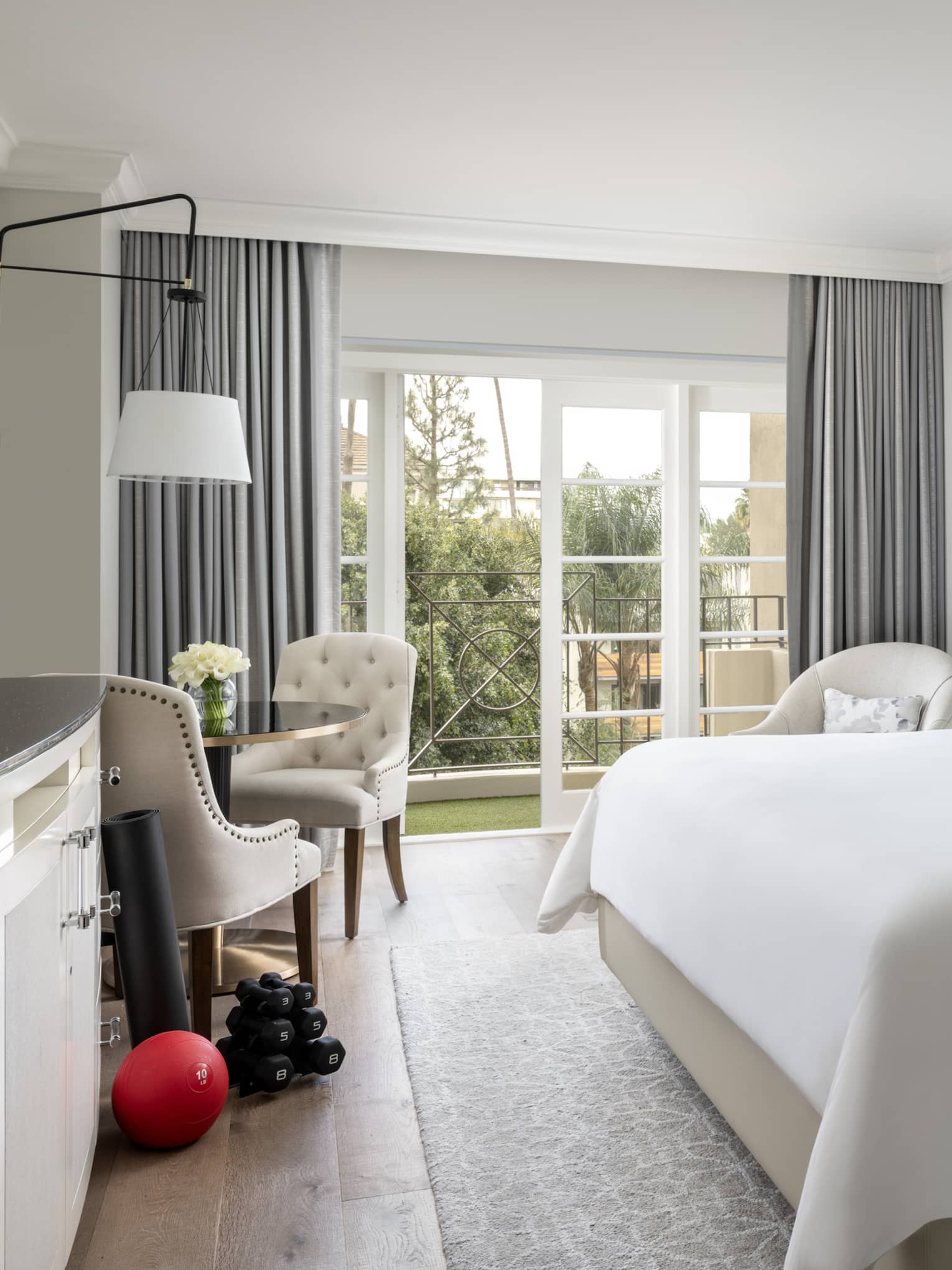 A hotel room with large open window, bed with white sheets, dresser, two chairs, an exercise ball and weights.