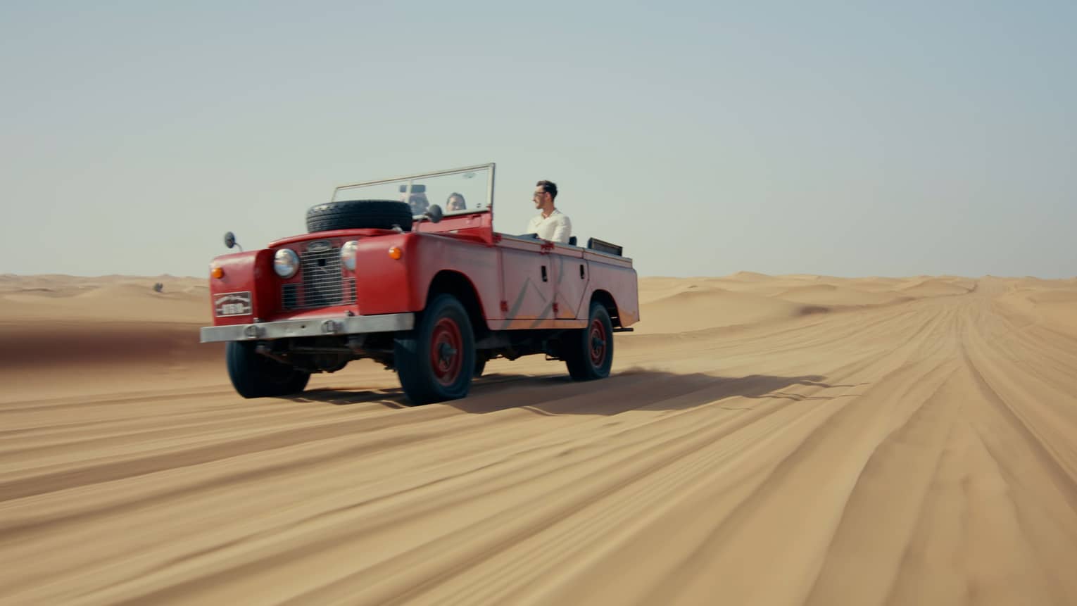 A red truck driving down a long stretch of sand under a blue sky.