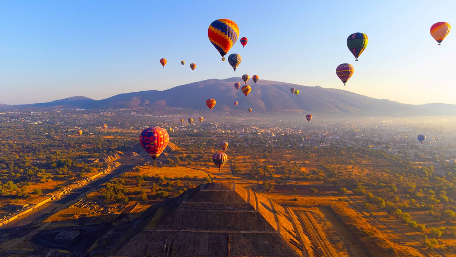 Hot air balloons floating over a pyramid structure with a mountain in the distance.