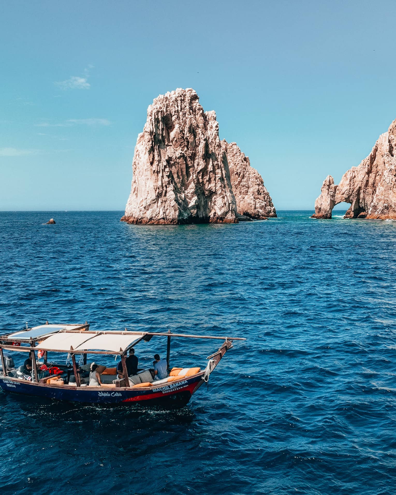A canopied wooden tour boat in open water with tall craggy rock formations jutting out of the ocean in the background.