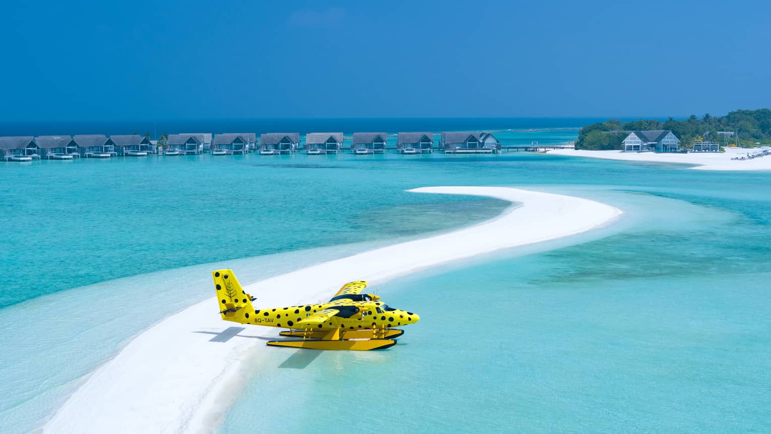 Yellow and black-spotted single-engine plane flies over clear waters of the Maldives, overwater bungalows in distance