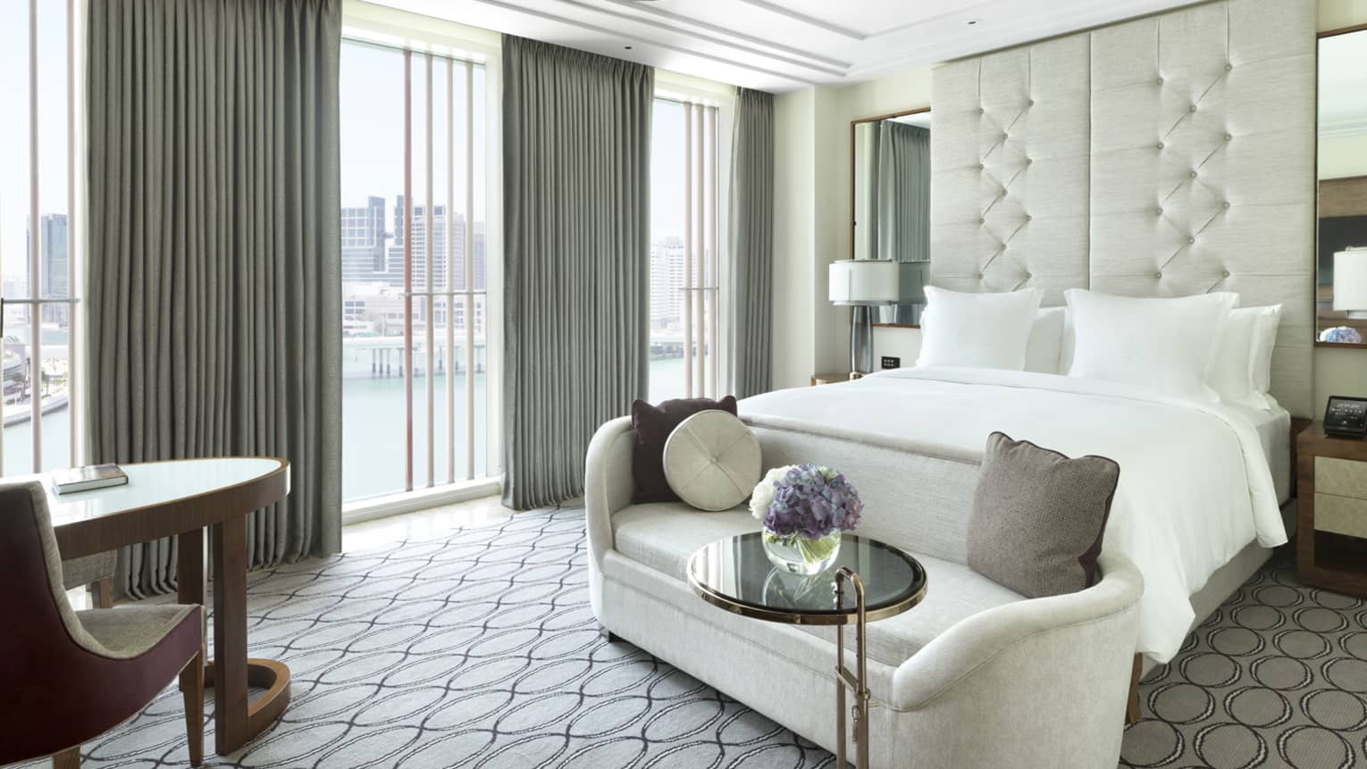 Deluxe room plush white loveseat at foot of bed by sunny window with grey curtains, table and chair