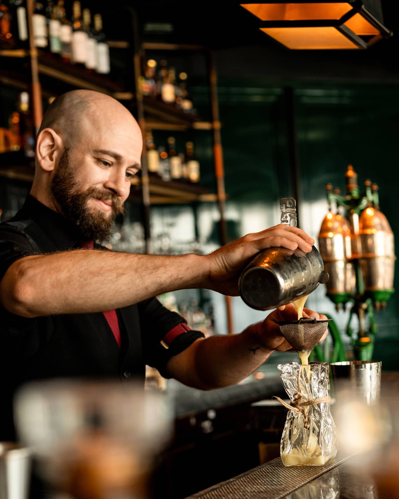 A smiling bartender pours a milky liquid through a strainer into a sculpted blown-glass tumbler, in a warm bar setting.